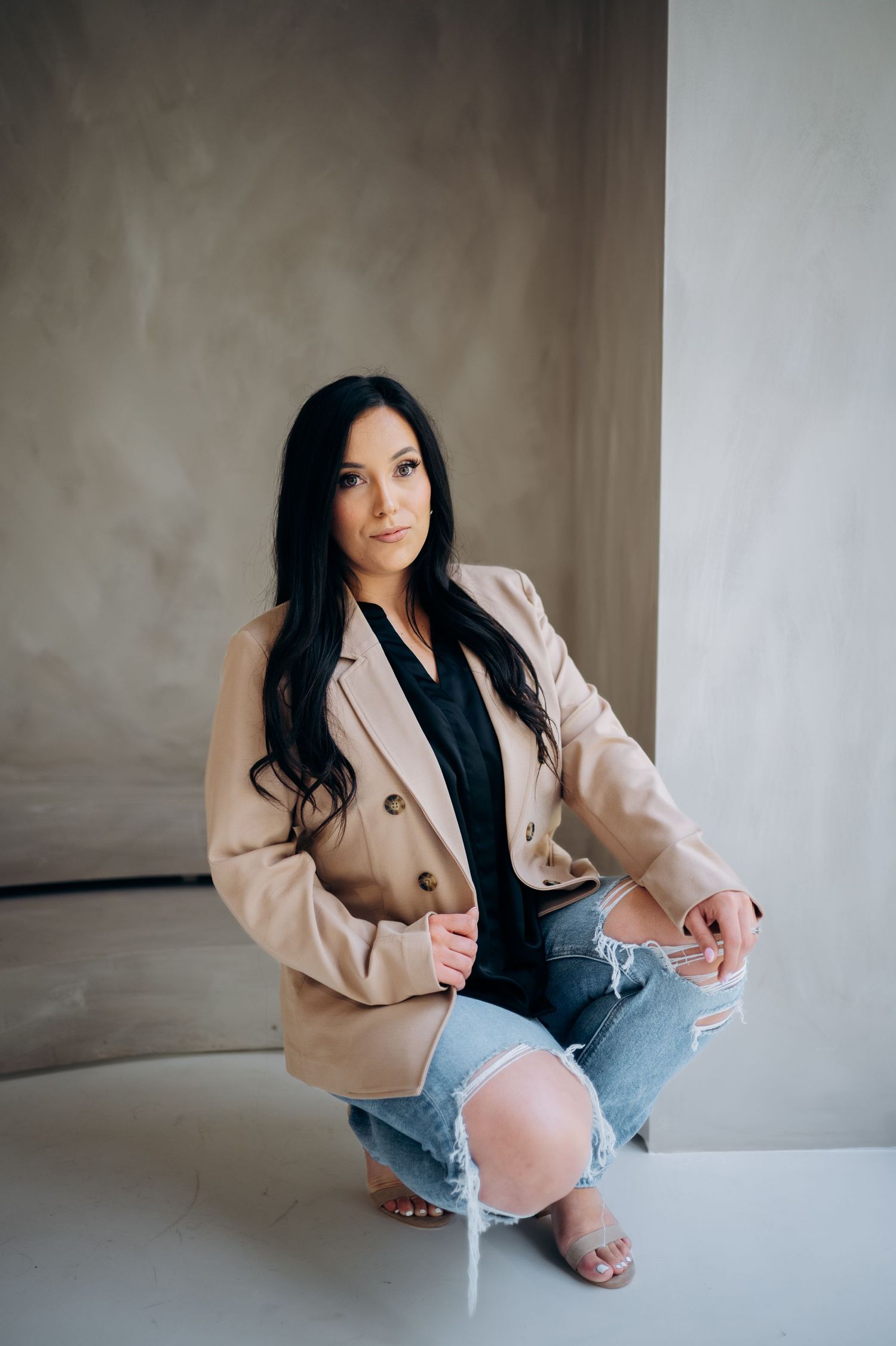 A fashion model poses in a beige blazer, black top and distressed jeans against a neutral wall.