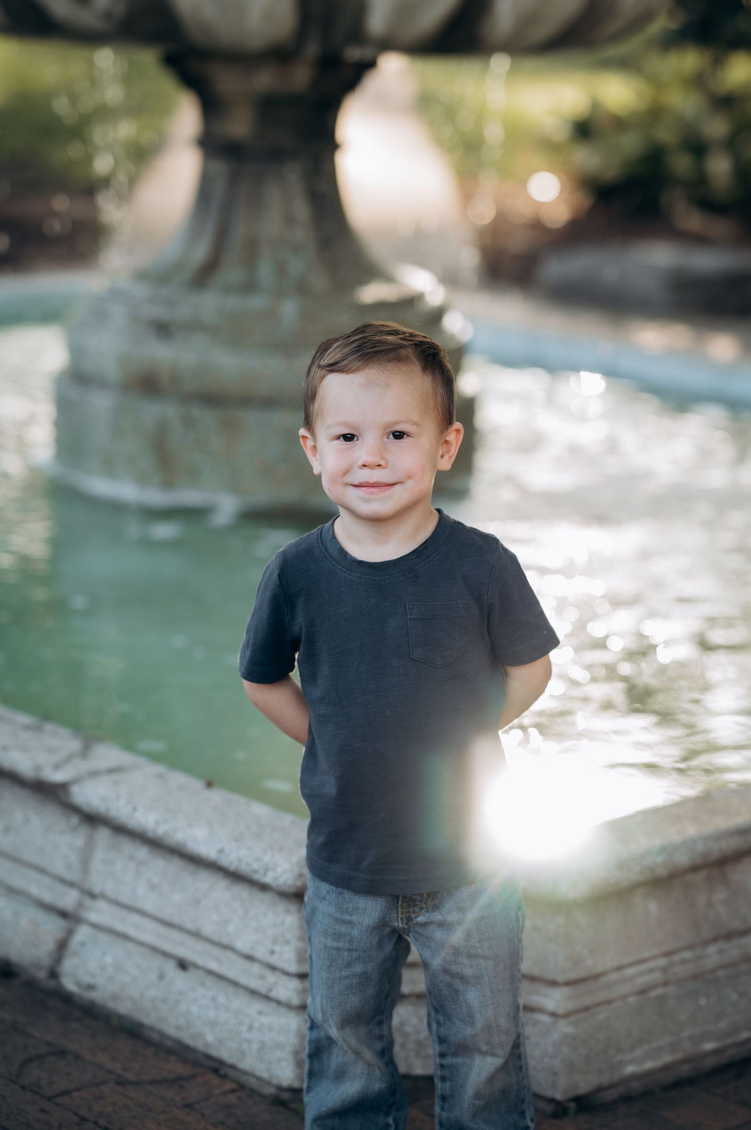 A young boy in a gray t-shirt and jeans smiles in front of a stone water fountain outdoors.
