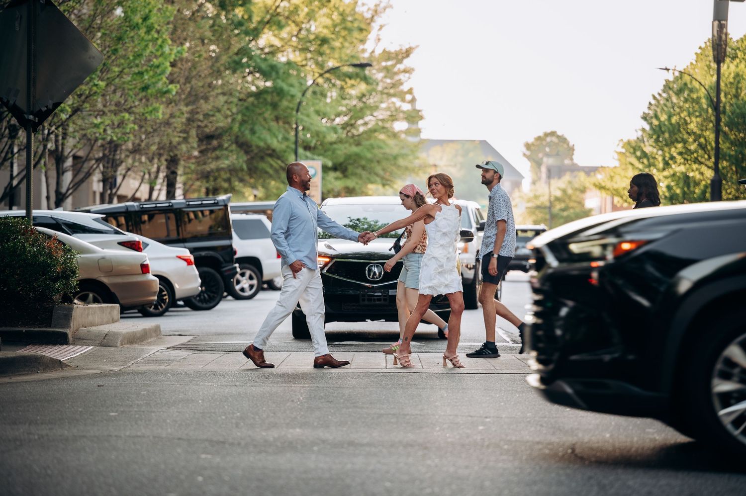Elegantly dressed couples walk through a sunlit outdoor shopping plaza with lush trees and parked cars.
