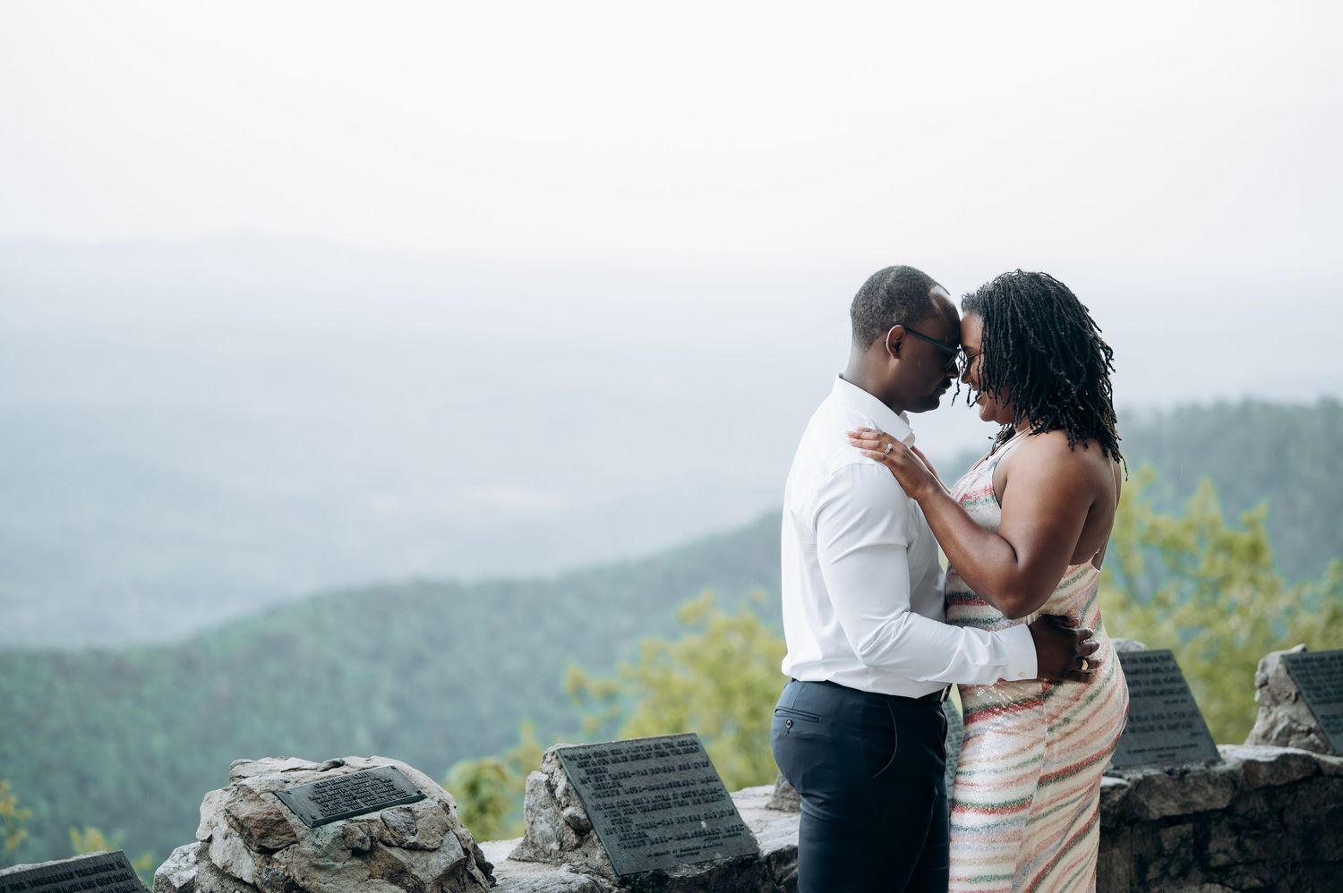 A couple embraces romantically on a stone overlook with misty mountain views in the background.