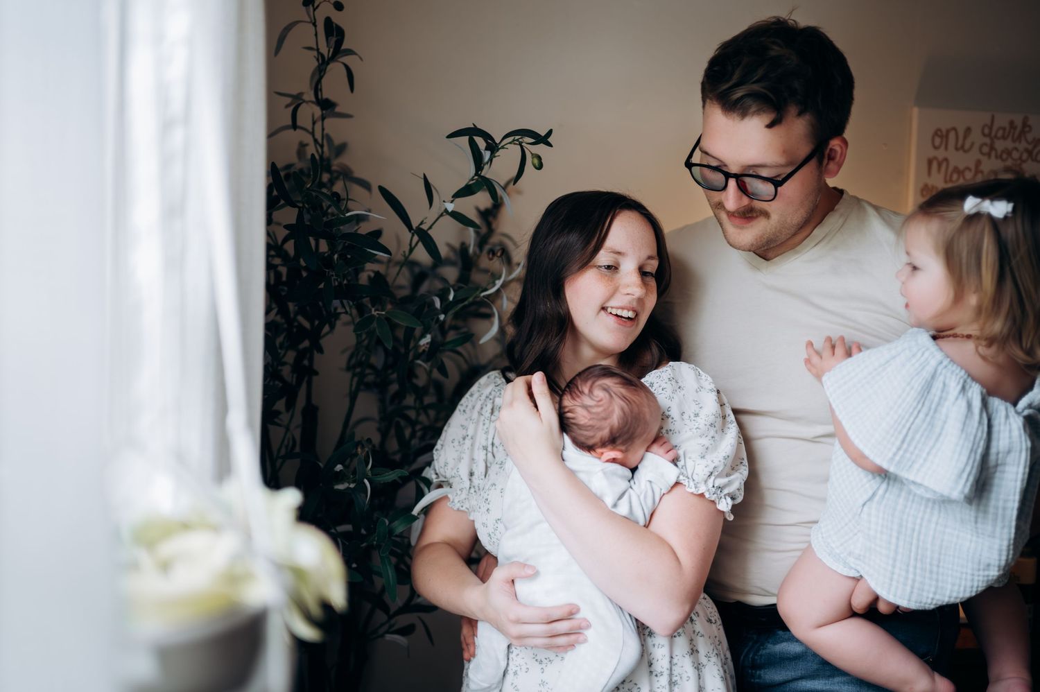 A happy family of four with a newborn baby gathered together by a window with green plants in a cozy home.