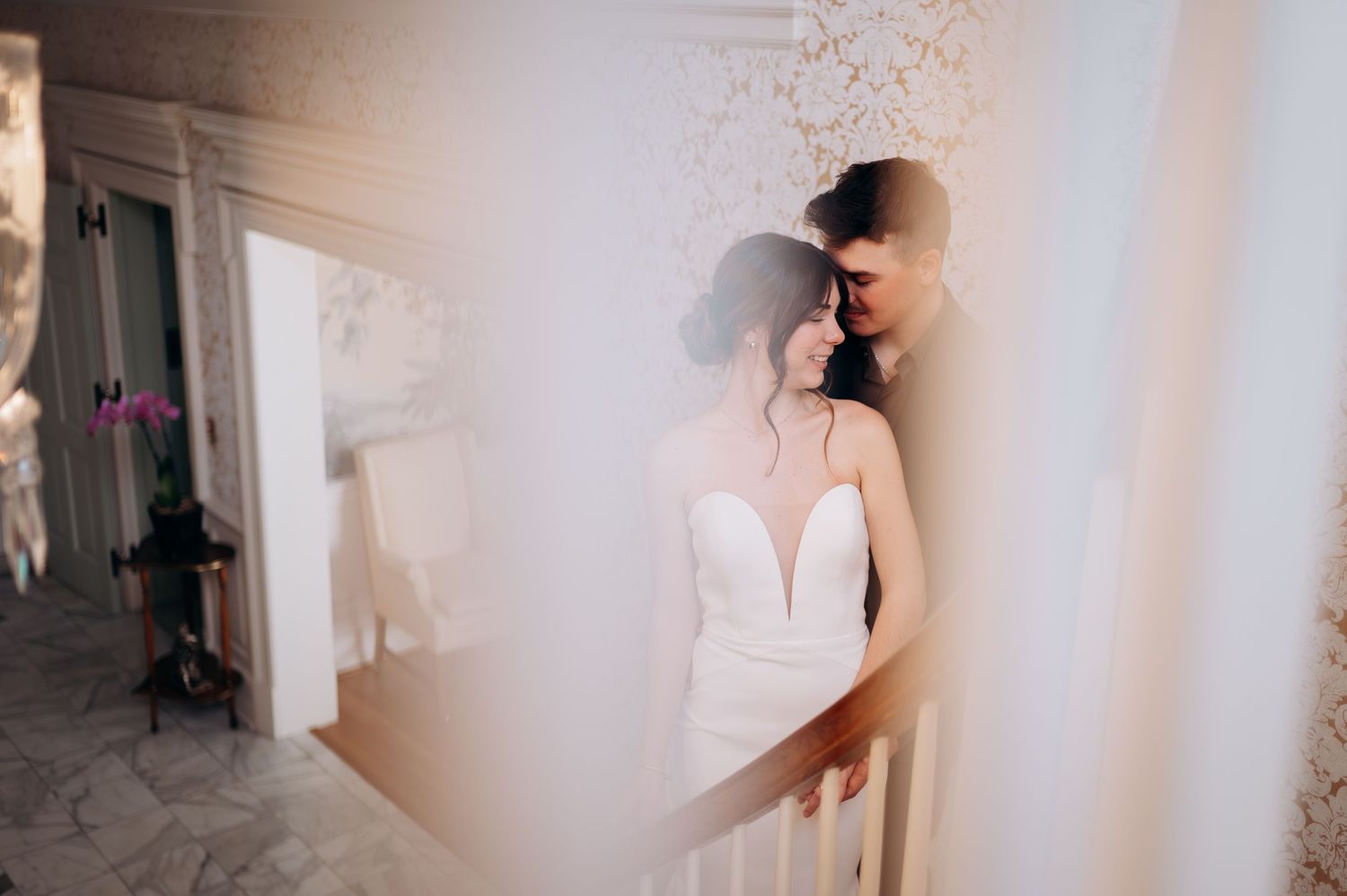 A bride in an elegant white wedding dress stands in soft window light creating a dreamy atmosphere.