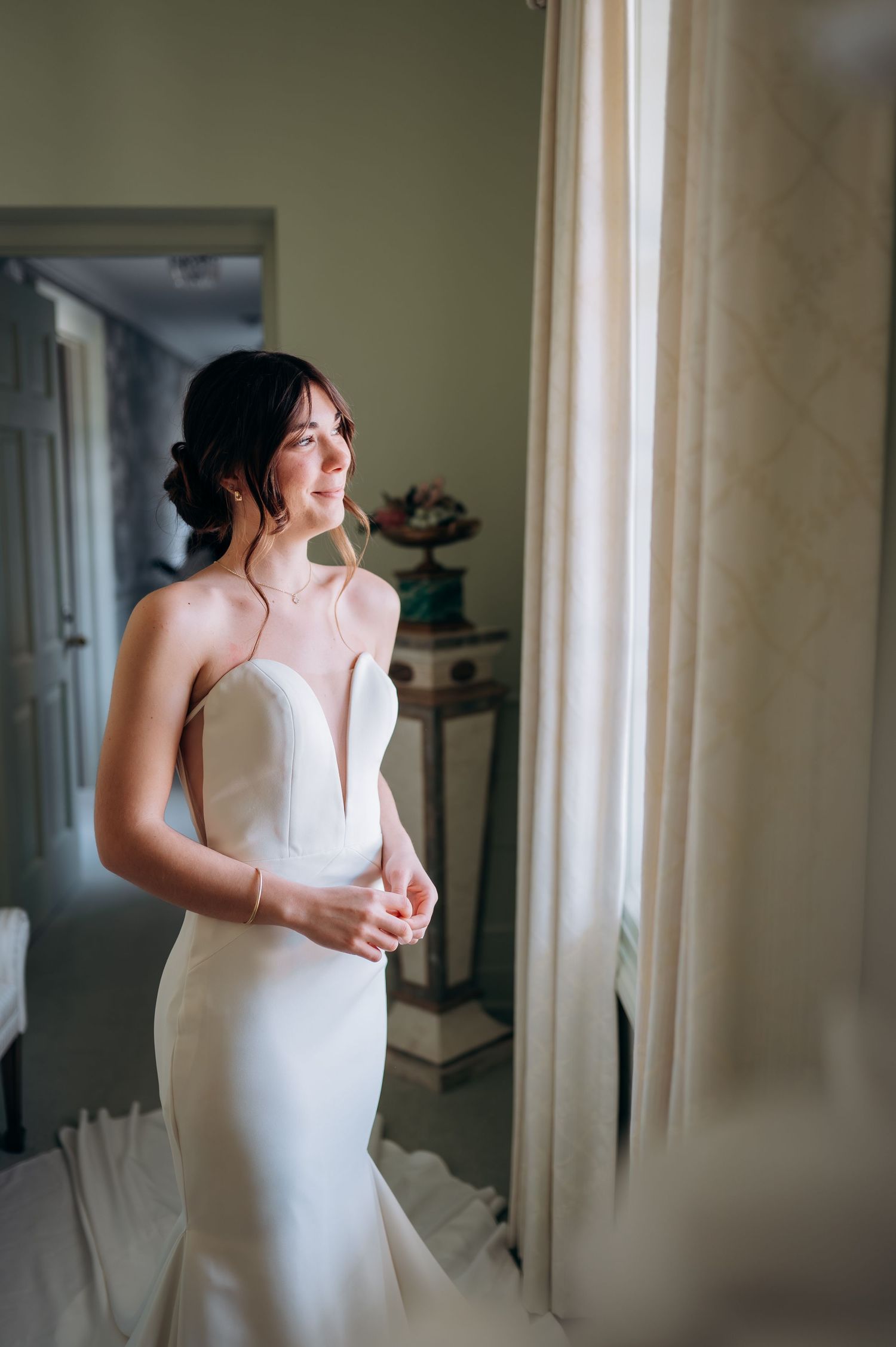 A bride in a sleek white gown gazes out a window in an elegant interior setting.