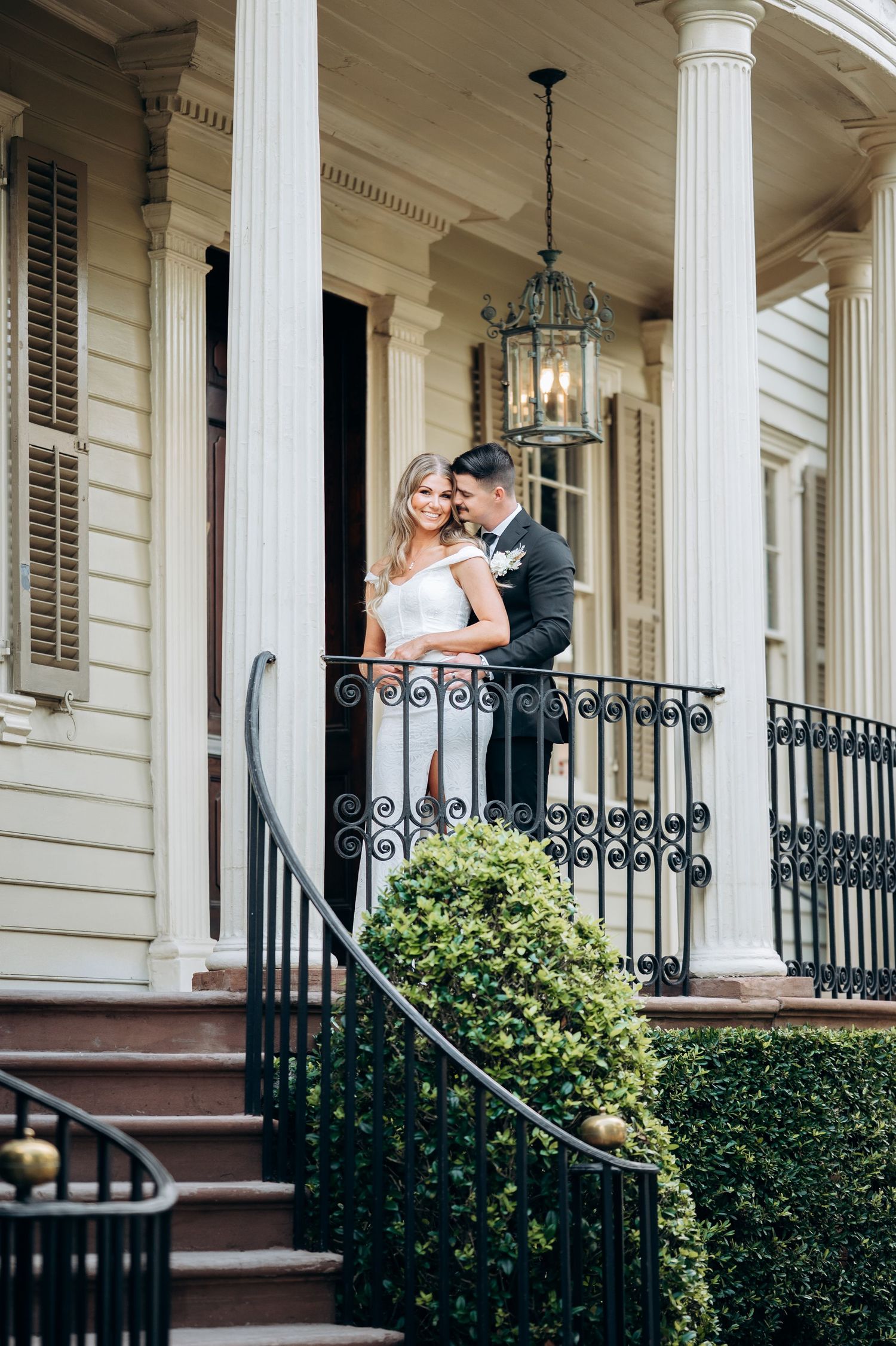 Bride and groom pose on elegant Southern mansion porch with wrought iron railing and lantern.