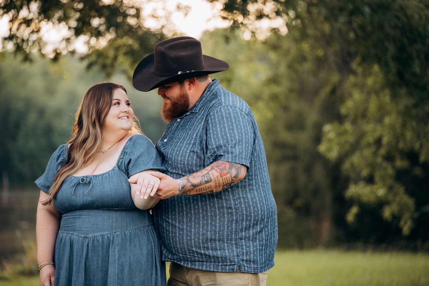 Couple smiling at each other outdoors, man in cowboy hat with tattoos holding hands with woman in blue dress.
