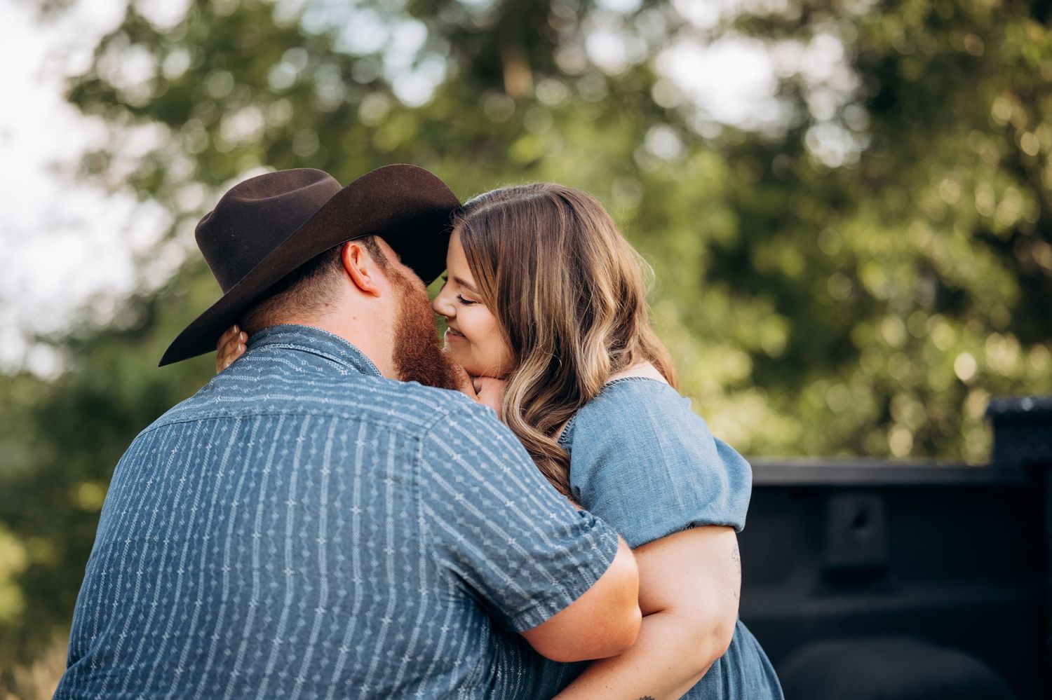 Couple sharing a tender kiss outdoors, man wearing a black cowboy hat and blue plaid shirt.