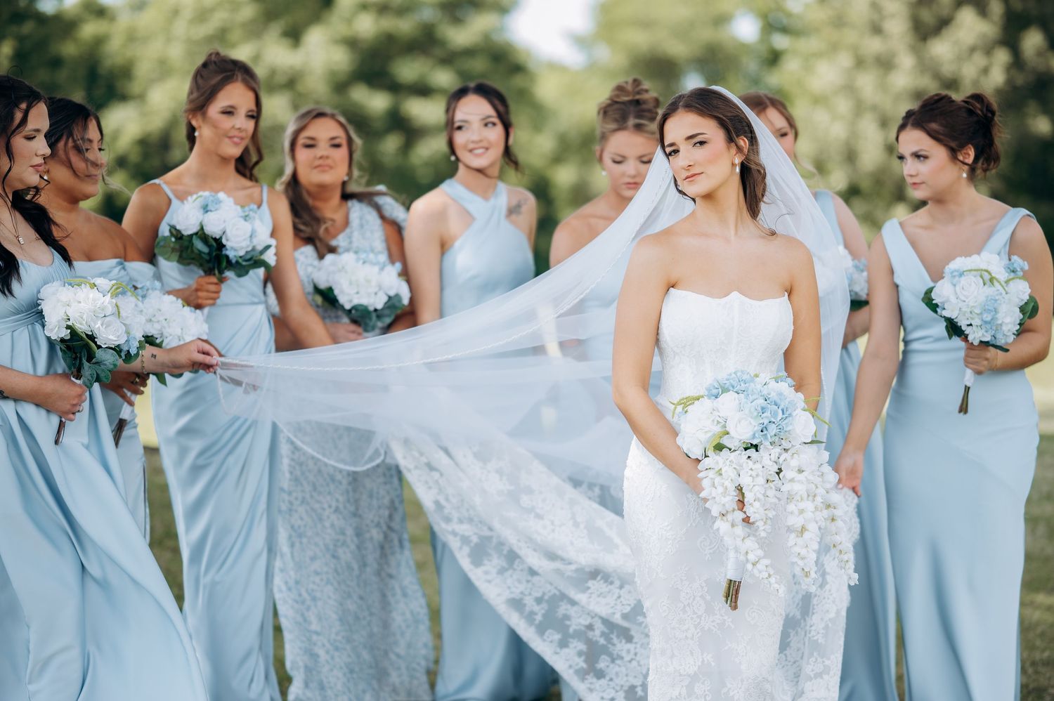Bride in white strapless gown holds bouquet while bridesmaids in light blue dresses hold flowing veil outdoors.