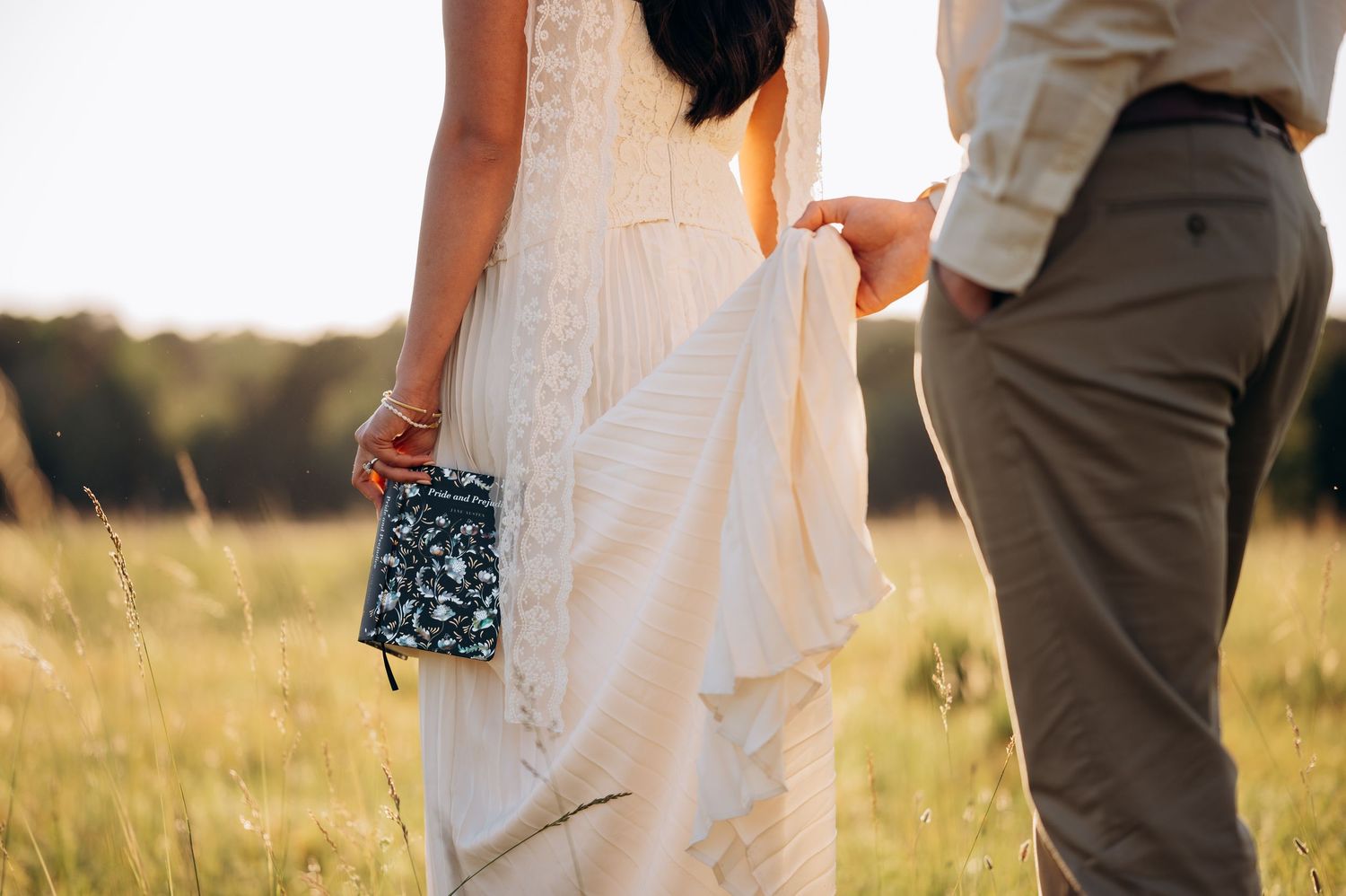 Bride in white lace gown holding clutch purse with groom in sunlit field at golden hour wedding.