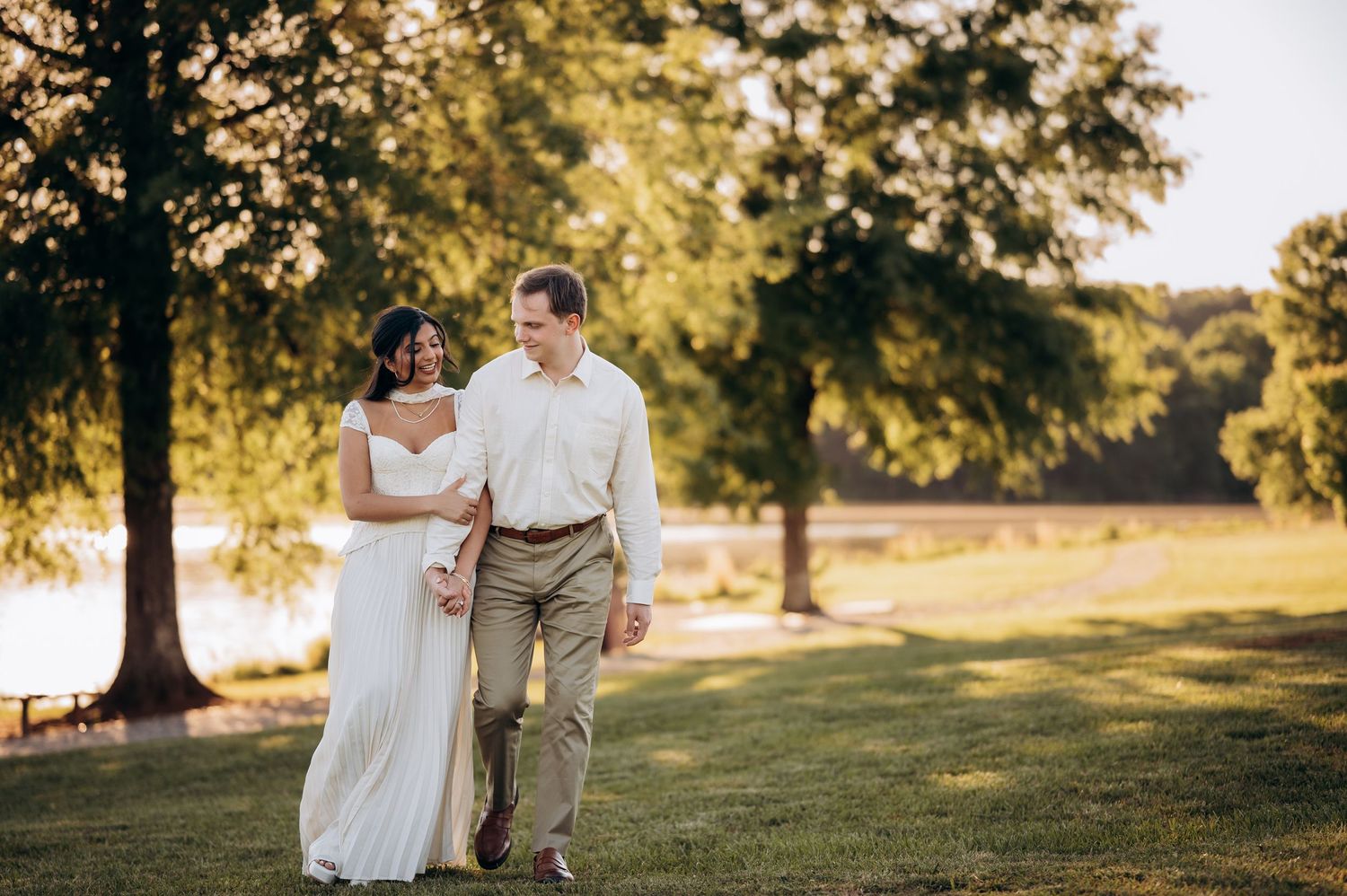 Newlywed couple walking hand in hand on green lawn near lake at golden hour, bride in white gown, groom in khaki.