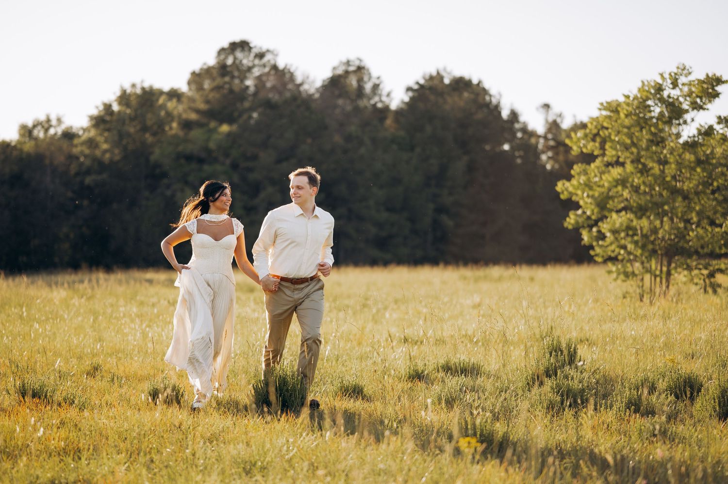 Bride and groom holding hands walking through a sunlit golden meadow with lush green trees in the background.