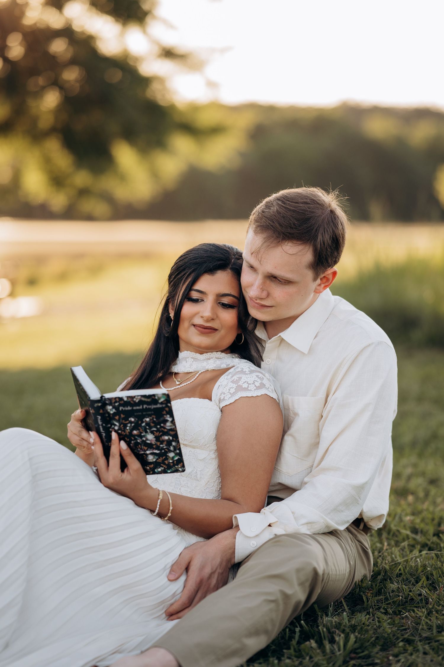 Couple in white attire sitting on grass, reading a book together near a lake at golden hour sunset.