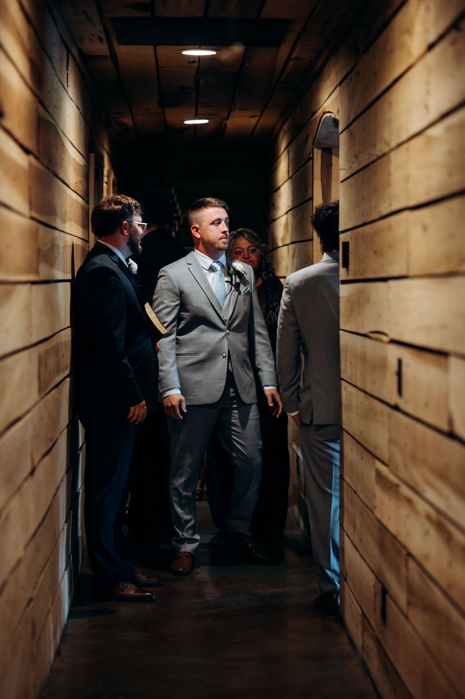 Groom in gray suit stands with groomsmen in a narrow wooden hallway before wedding ceremony.