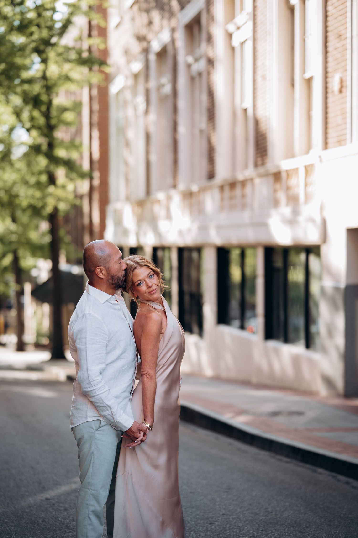 Couple embracing tenderly on a sunny urban street, man in white shirt and grey pants, woman in satin slip dress.