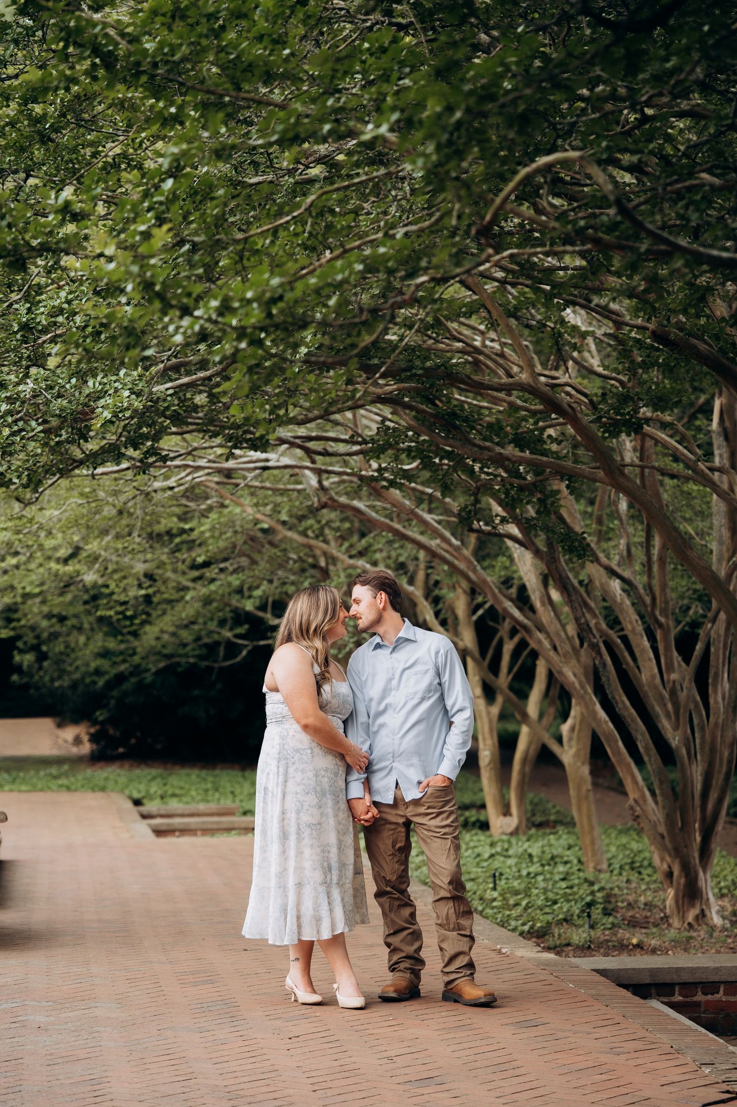 Couple sharing a tender moment on brick walkway under canopy of trees during engagement session.