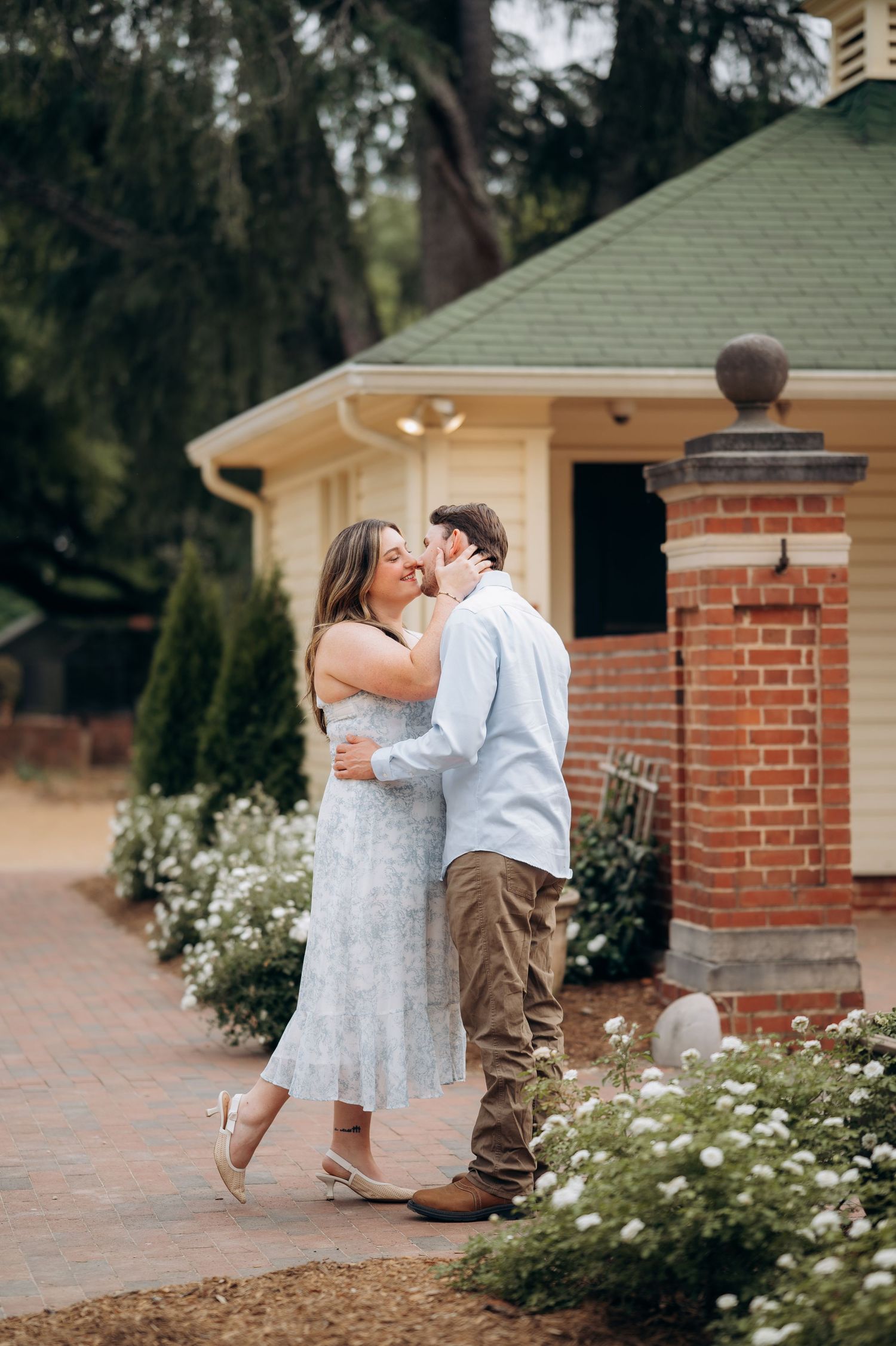 Couple sharing a romantic kiss near brick building with white flowers during outdoor engagement photos.