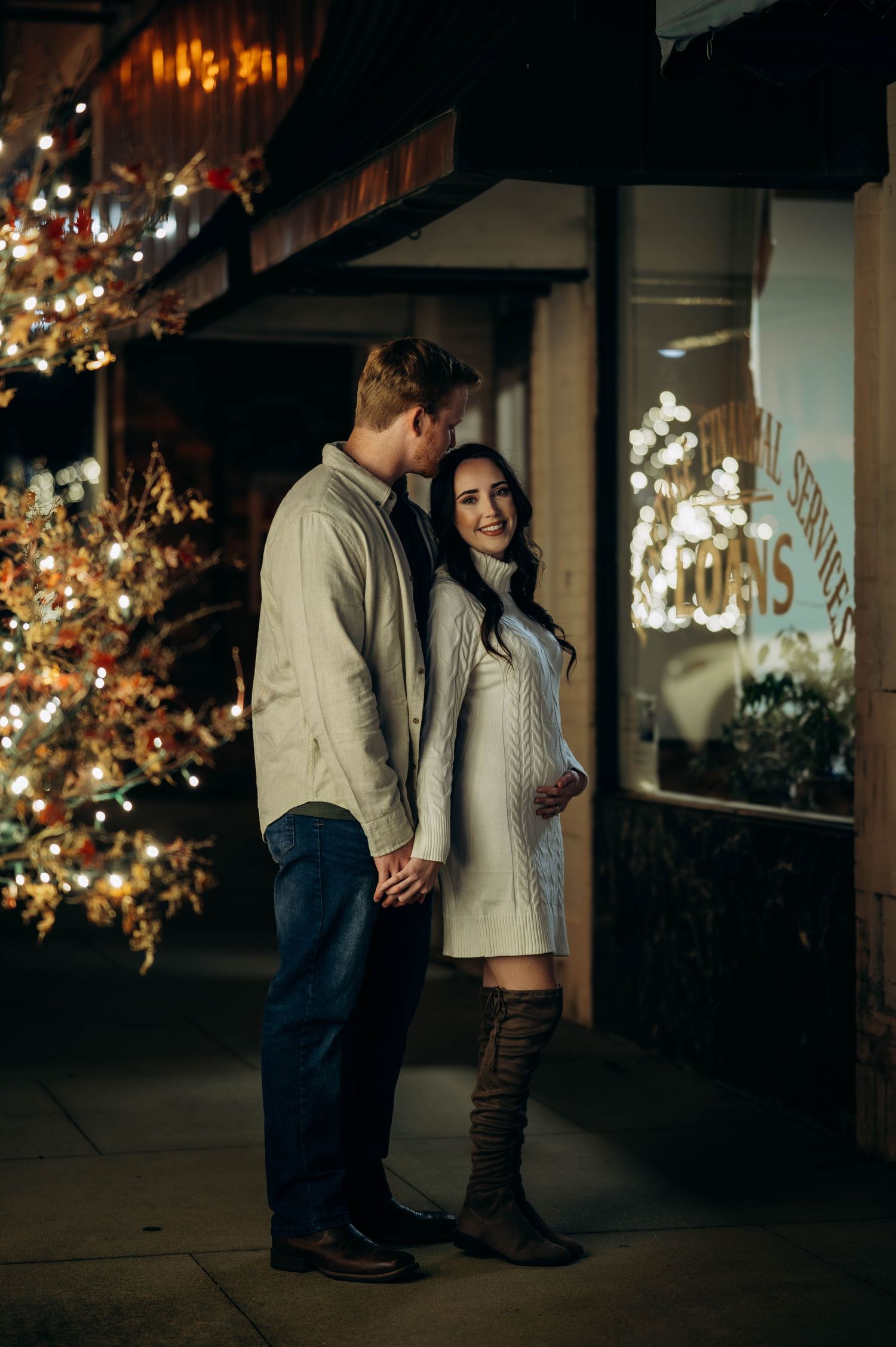 Couple sharing tender moment outside festive storefront with Christmas lights at night.