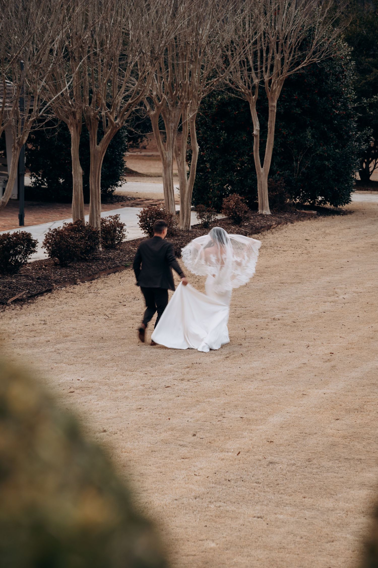 Bride in flowing white gown and veil running with groom through tree-lined garden path.