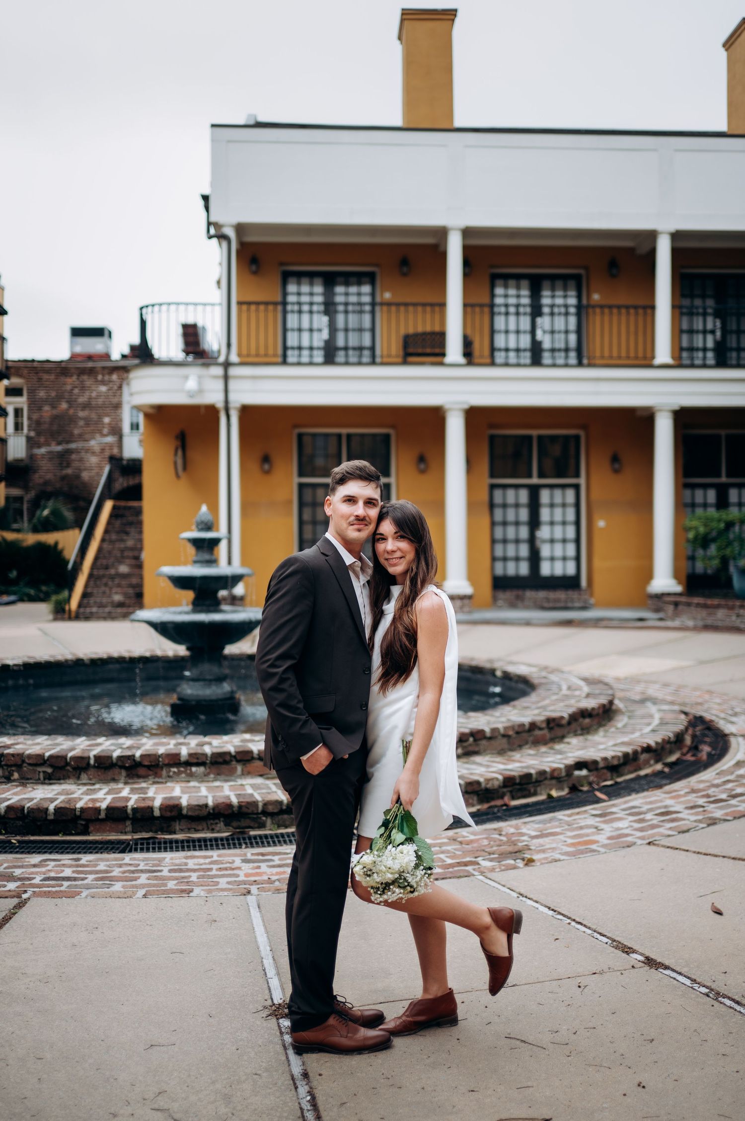 Couple poses in front of yellow building with fountain during wedding day in New Orleans.