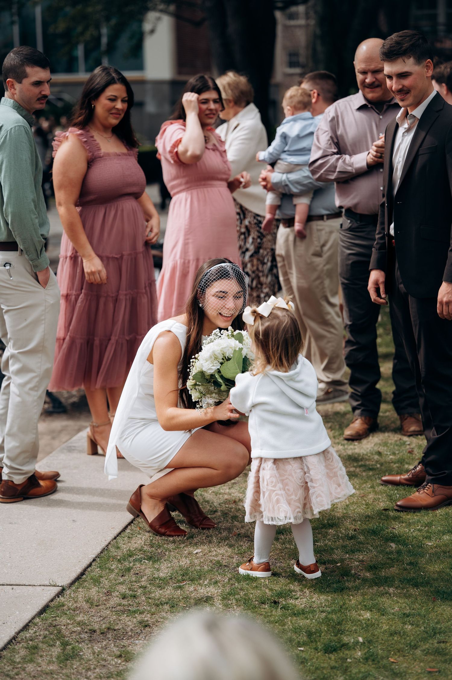 Bride in white dress crouches to greet a small flower girl holding a bouquet at outdoor wedding.