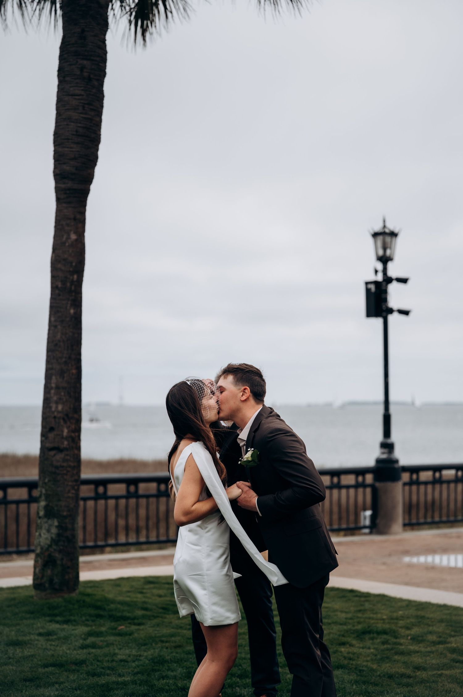 Newlywed couple sharing first kiss near palm tree with ocean and street lamp in background.