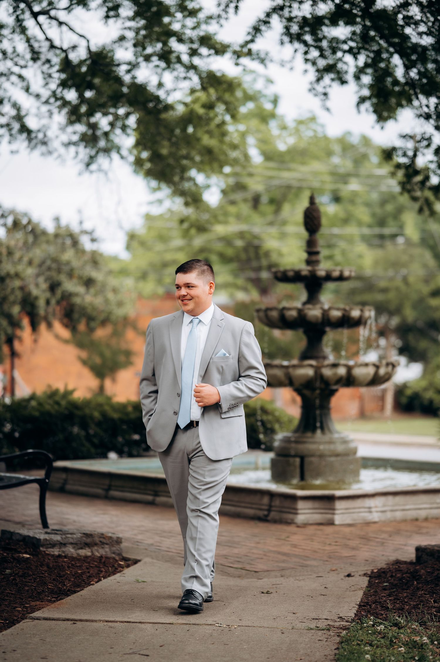 Smiling groom in light gray suit walking near ornate stone fountain in lush outdoor garden setting.