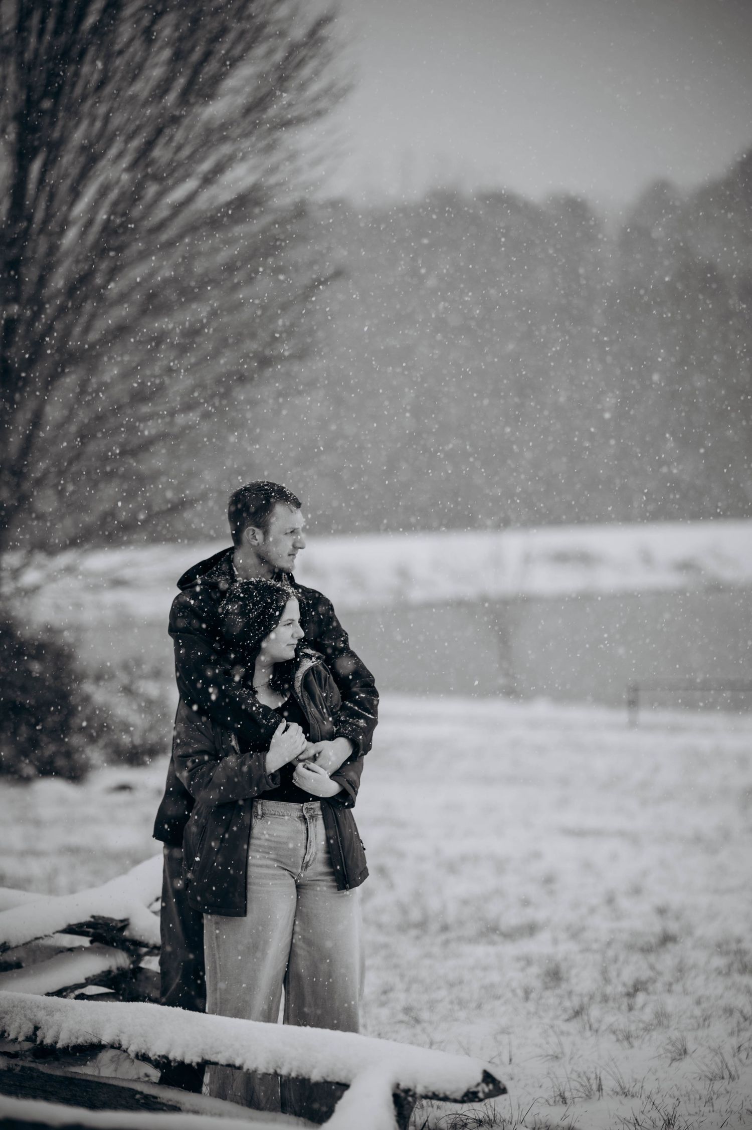 A couple stands close together in a snowy winter landscape with bare trees and a fence visible.