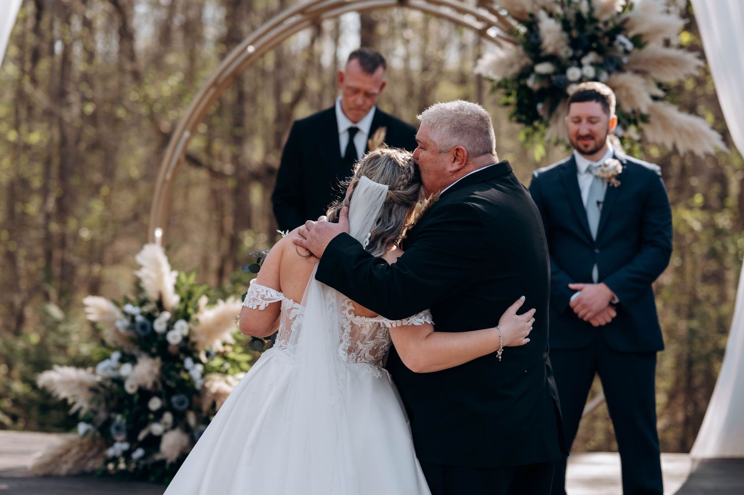 Wedding ceremony under floral arch with white draping in forest setting.