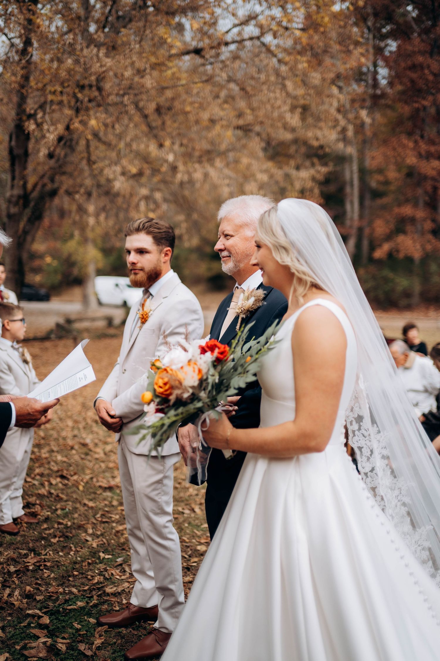 Wedding party interacts during outdoor fall ceremony with white dresses and light gray suits.