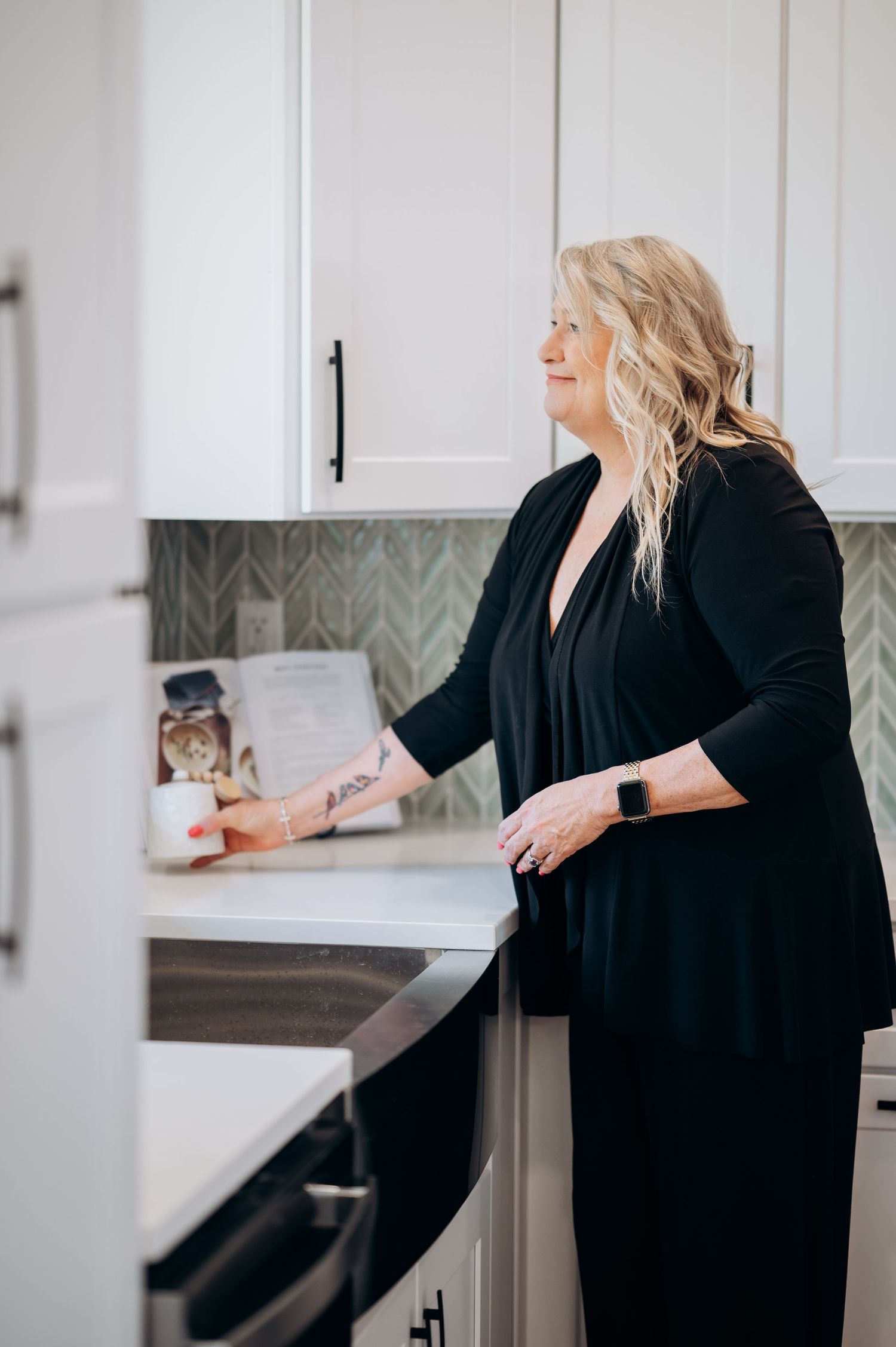 Blonde woman in black dress standing in modern white kitchen with green tile backsplash and smart watch.