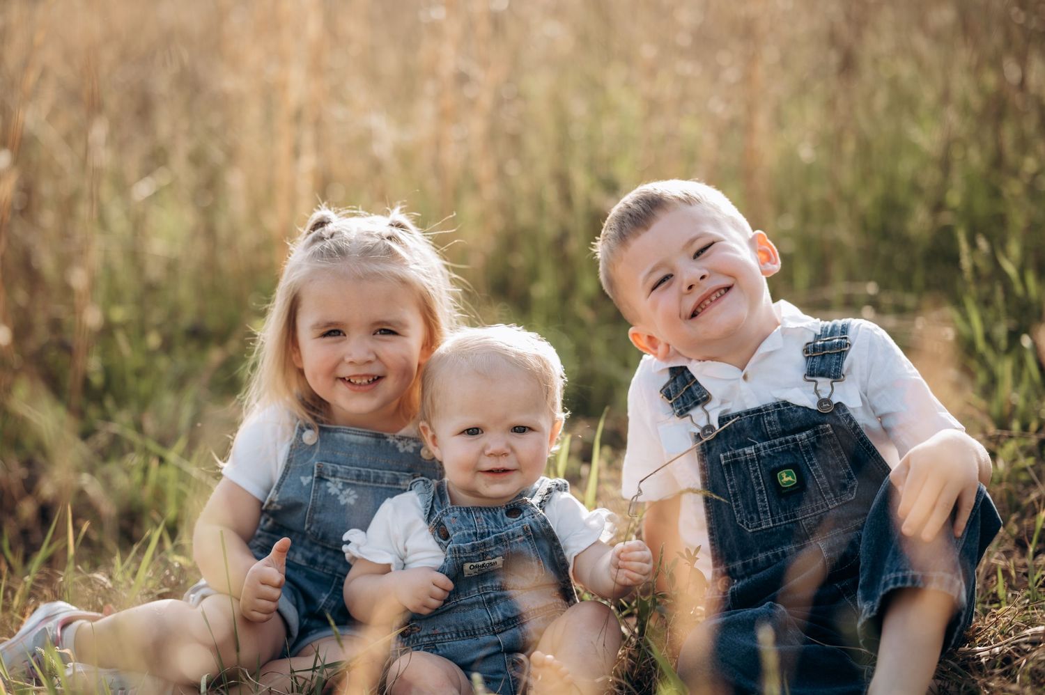 Three young siblings in denim overalls sitting together in a sunny field, smiling happily for a family portrait.