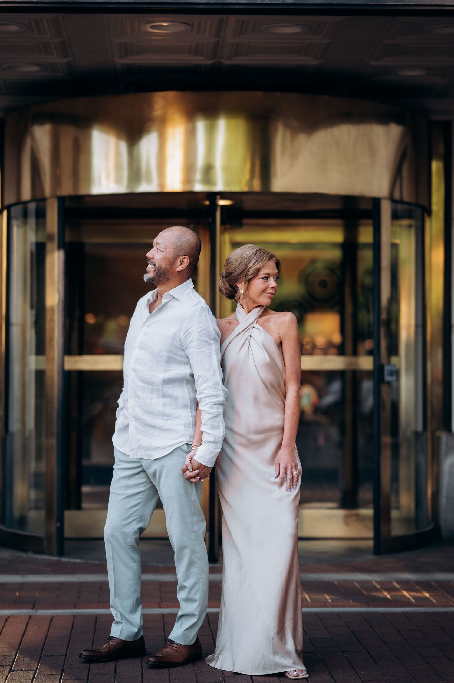 Stylish couple standing back-to-back holding hands before a golden revolving door, dressed in elegant semi-formal attire.
