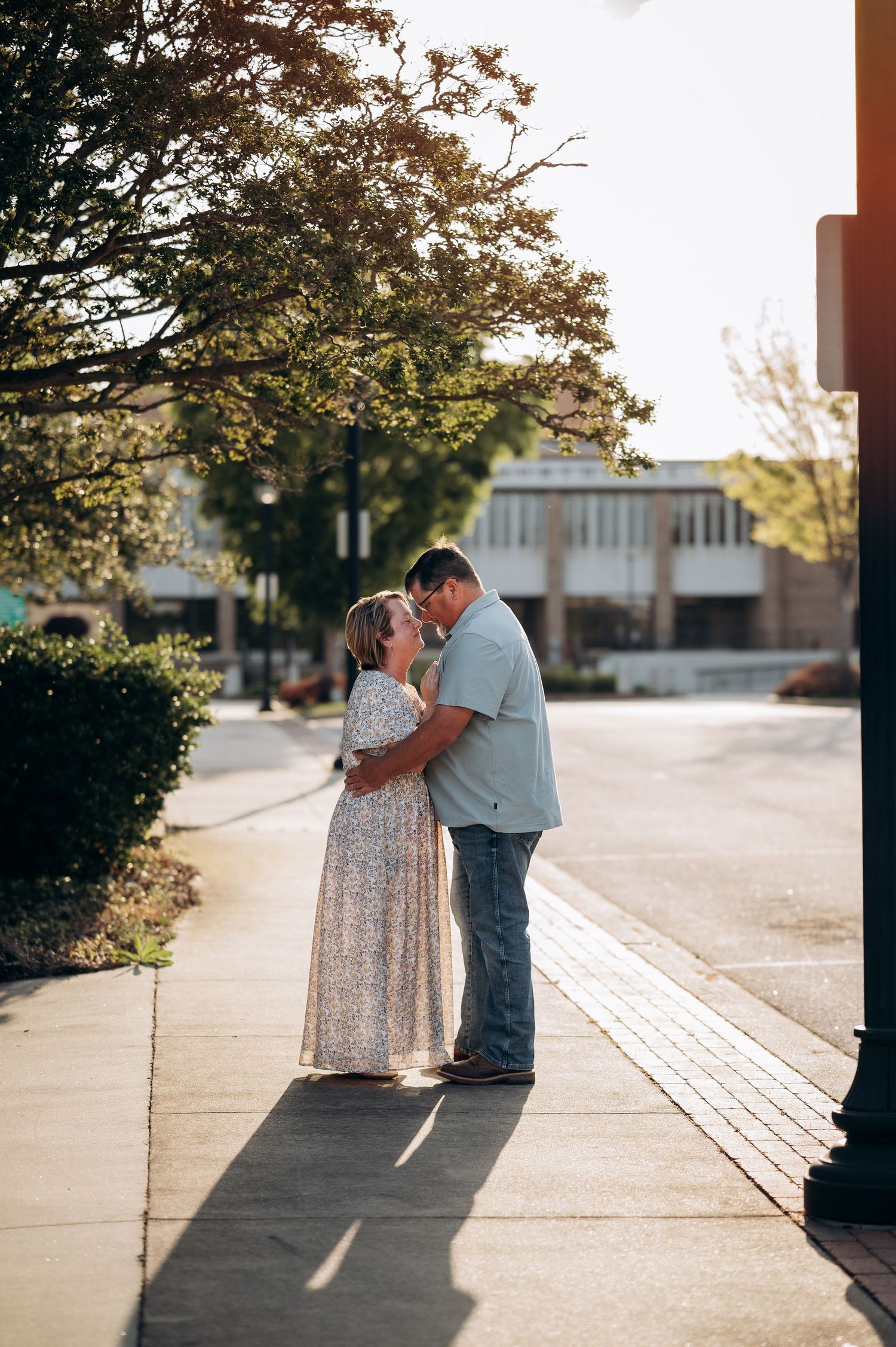 Couple sharing a romantic kiss on a sunny sidewalk surrounded by lush green trees.