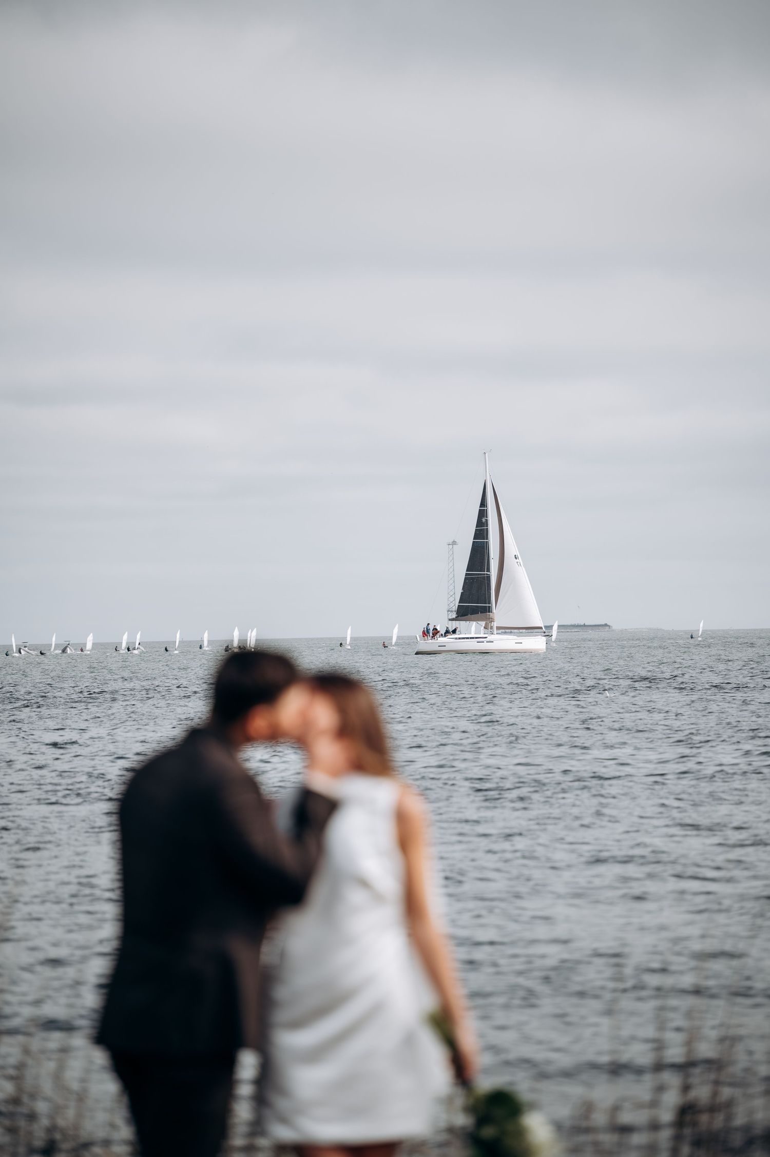 Couple embracing by waterfront with sailboats visible in background on overcast day.