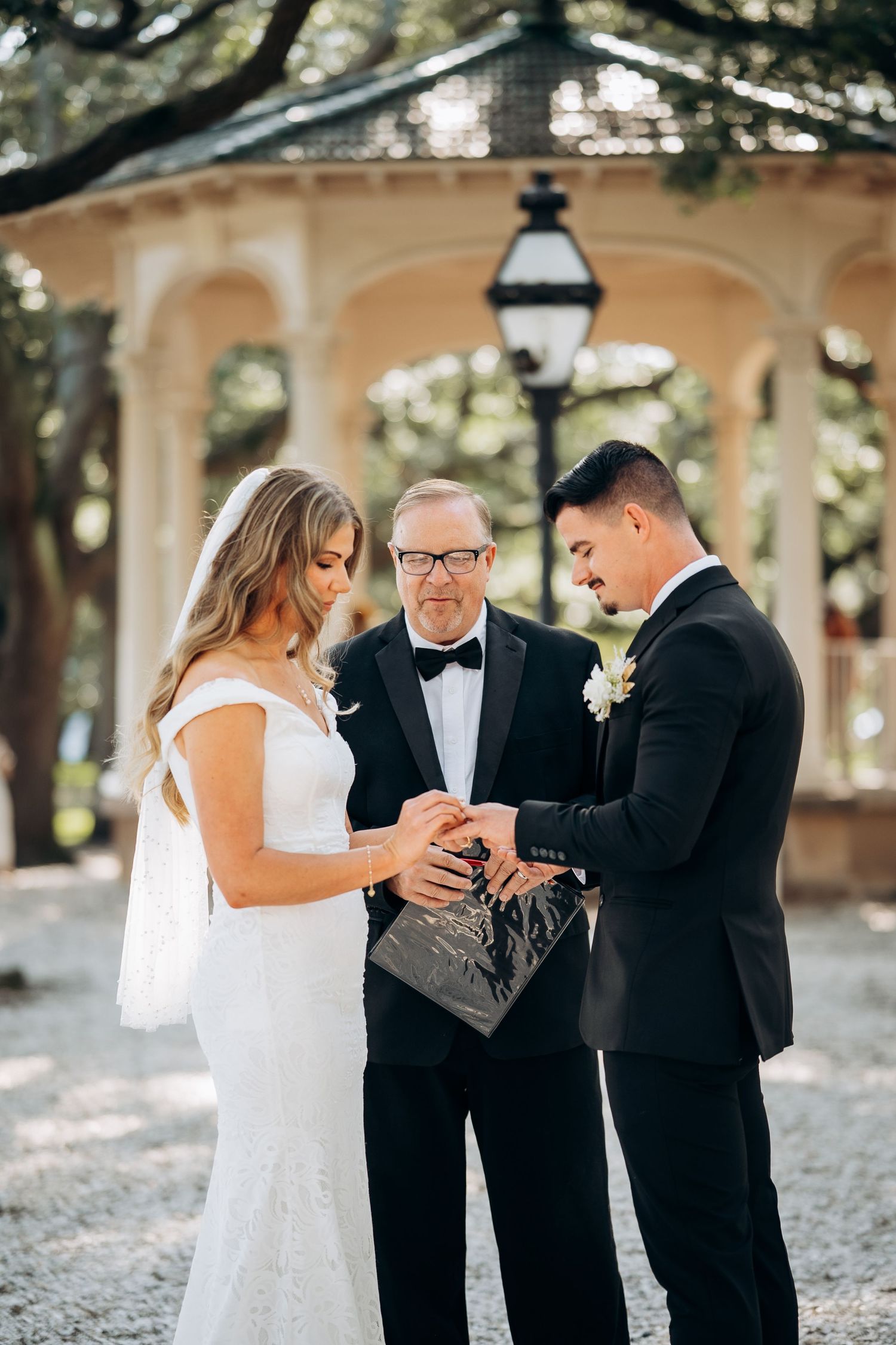 Bride and groom exchange rings with officiant at elegant gazebo outdoor wedding ceremony.