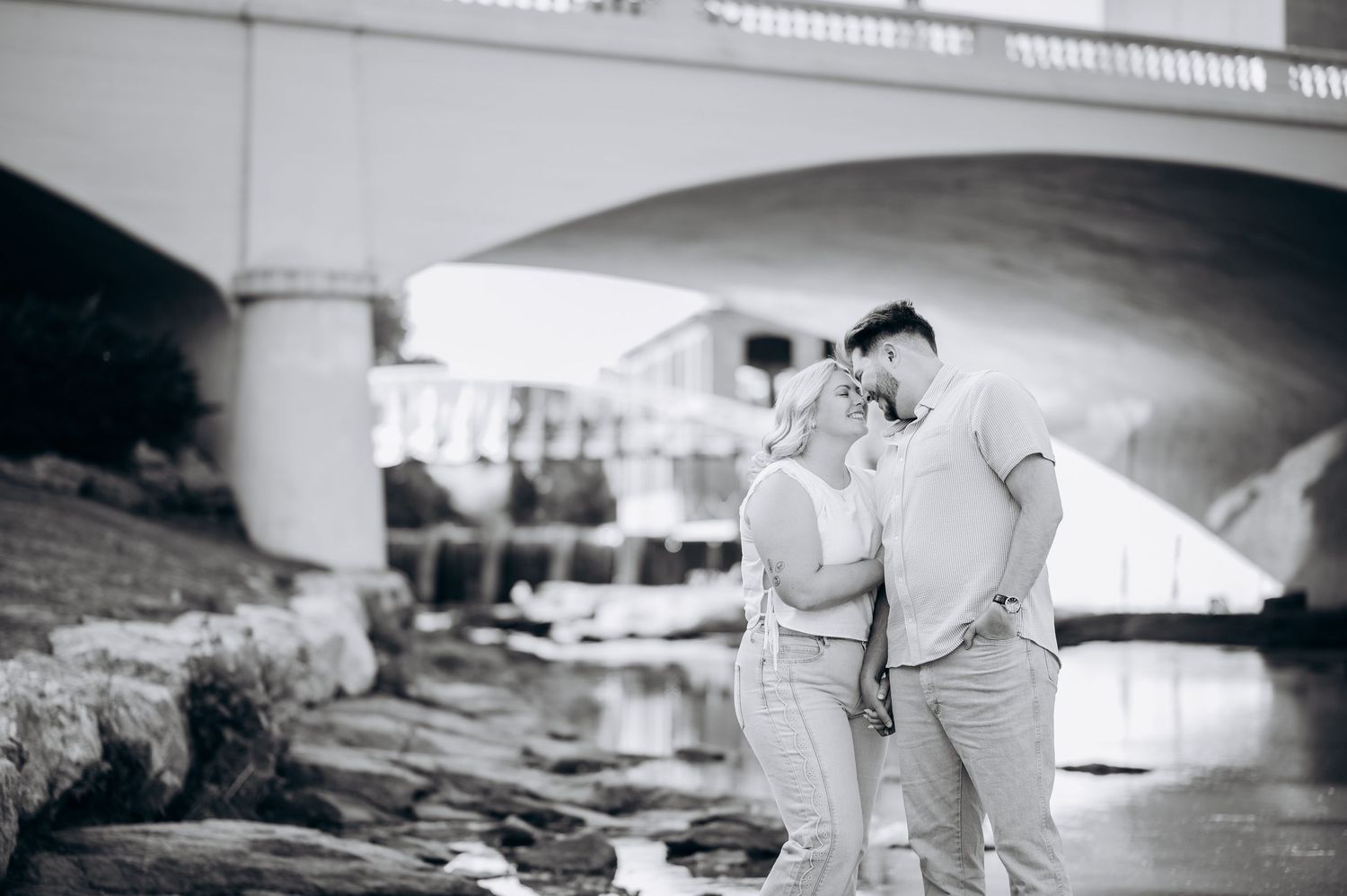 Black and white photo of a couple embracing near a rocky riverbank with an arched bridge in the background.