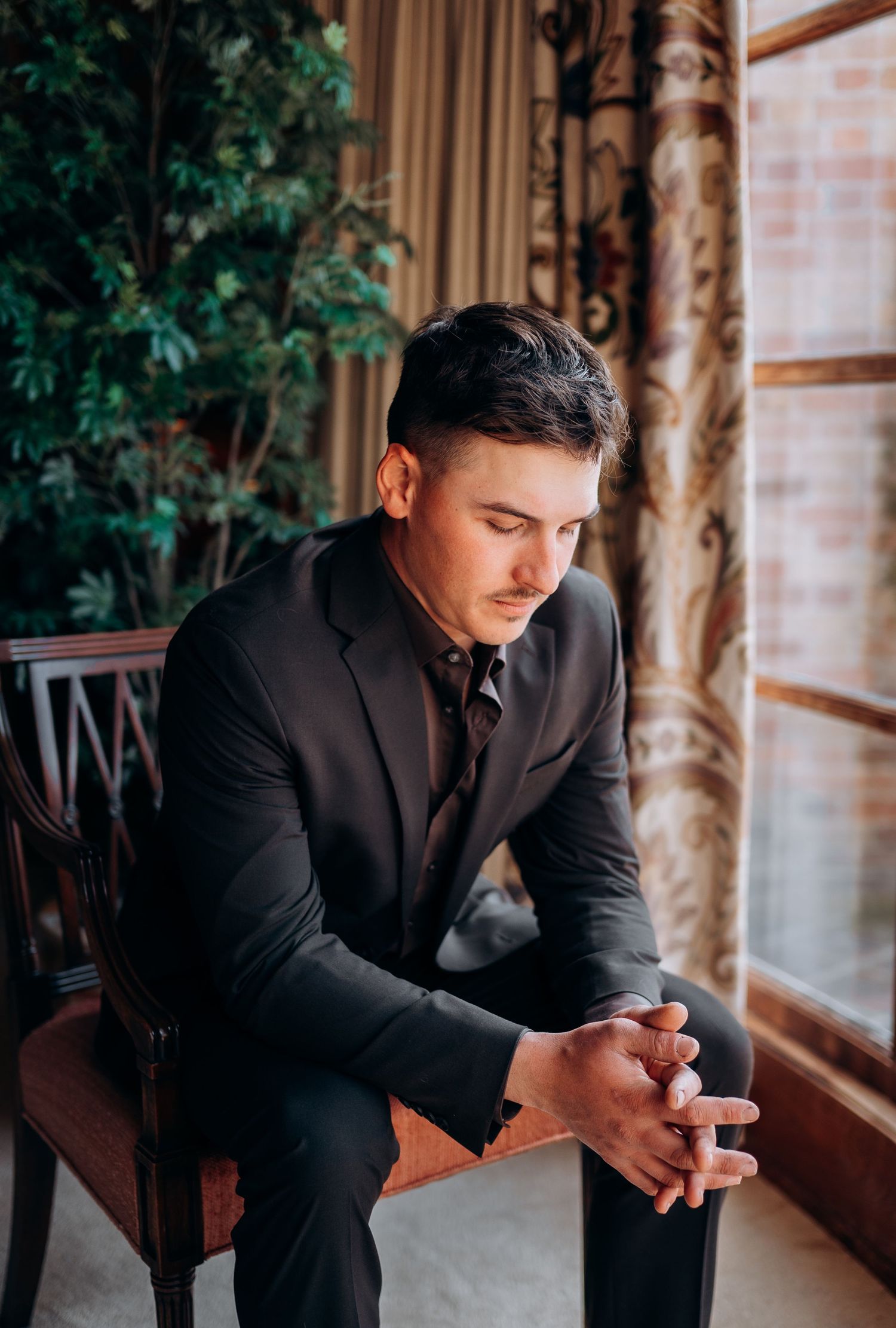 Person in dark suit sits by window checking phone in elegant room with floral curtains.