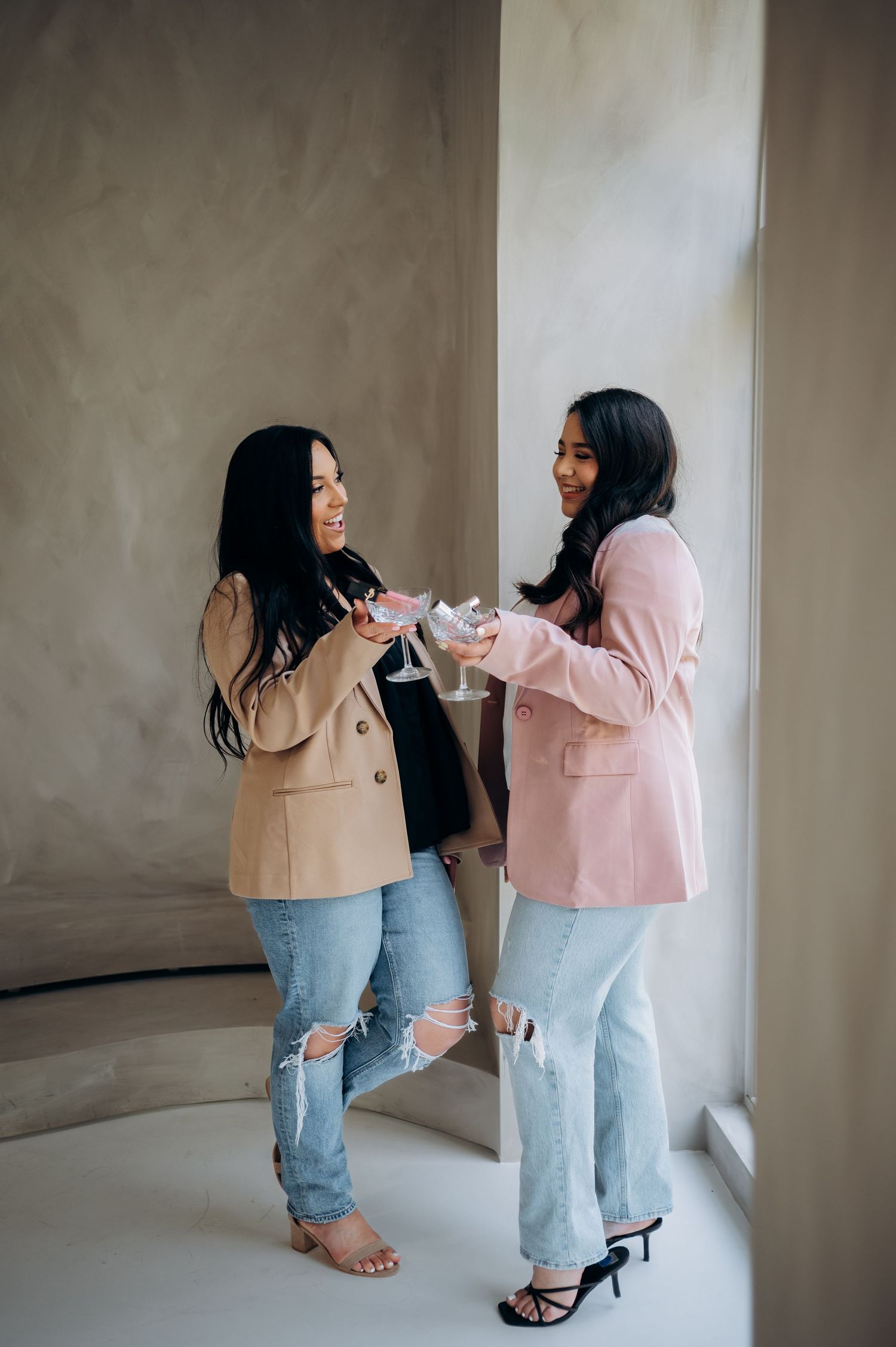 Two women in matching beige and pink blazers pose together against a neutral wall in a series of candid shots.