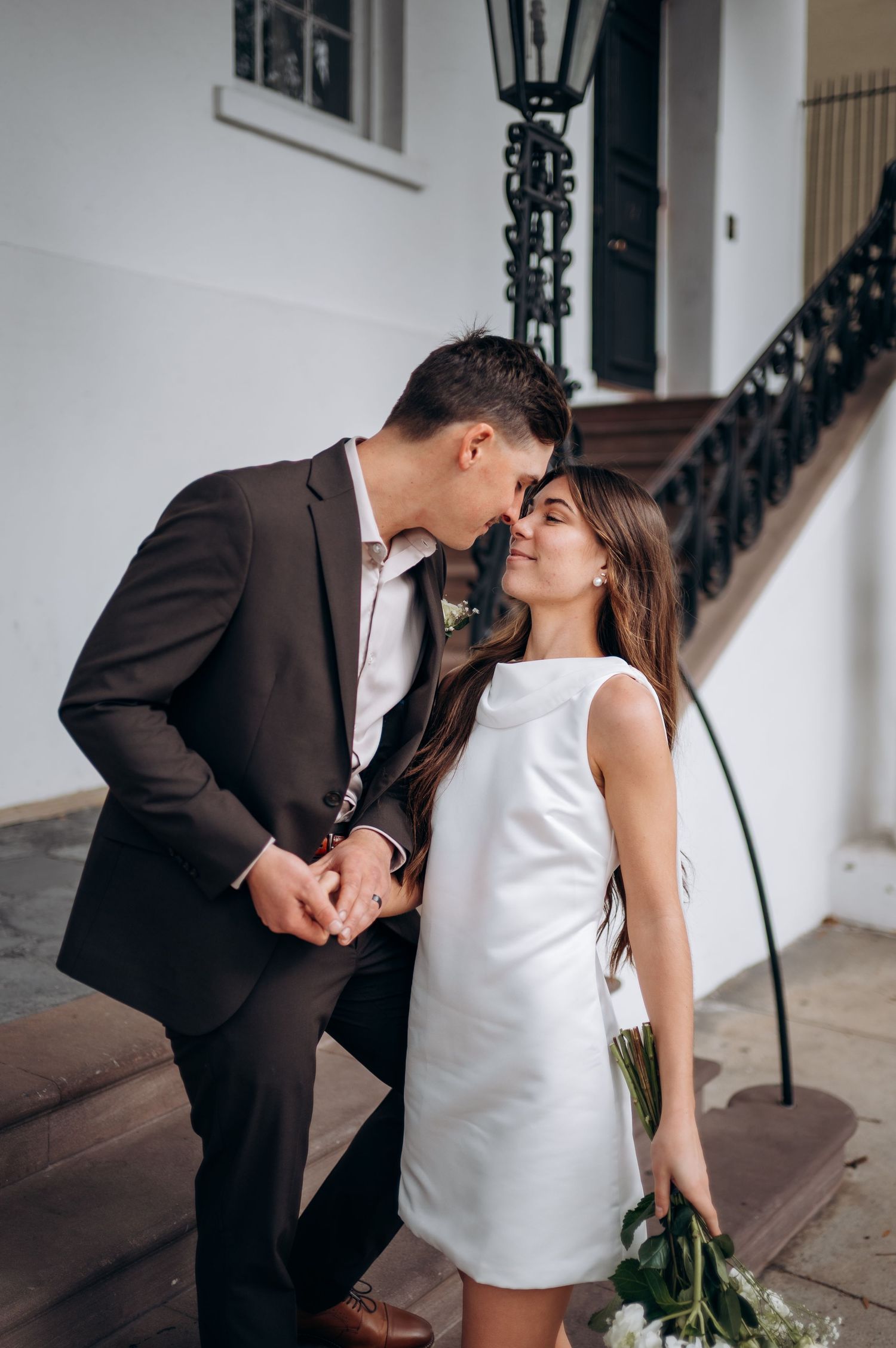 A romantic moment between a couple in formal wear standing by an ornate staircase with white flowers.