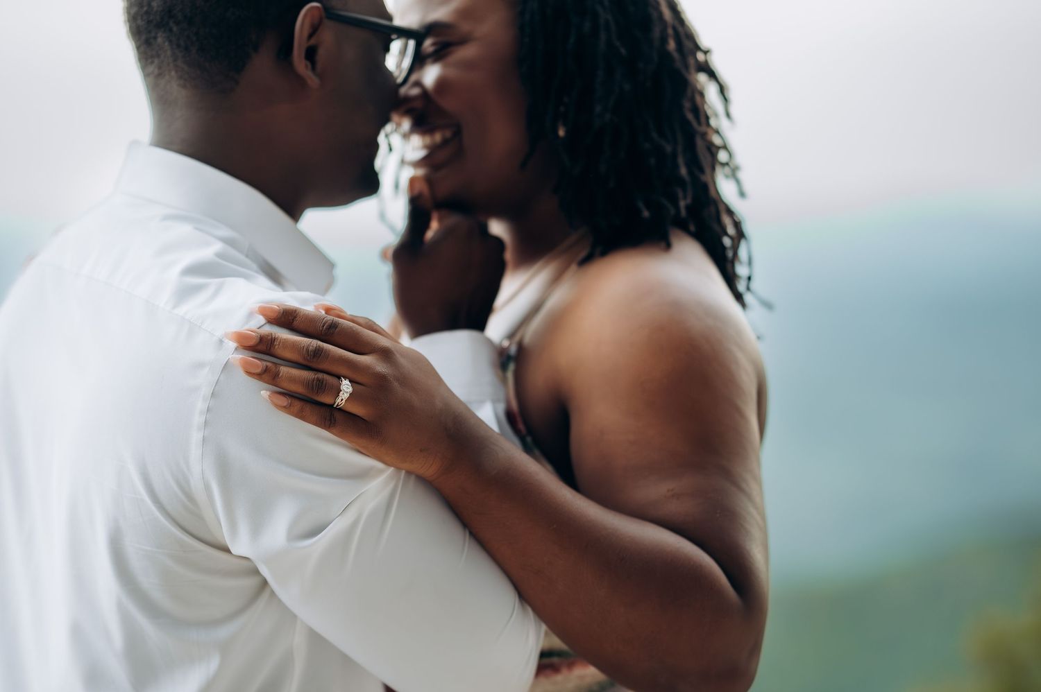 A joyful Black couple shares an intimate moment, the bride wearing a ring and white dress, embracing outdoors.