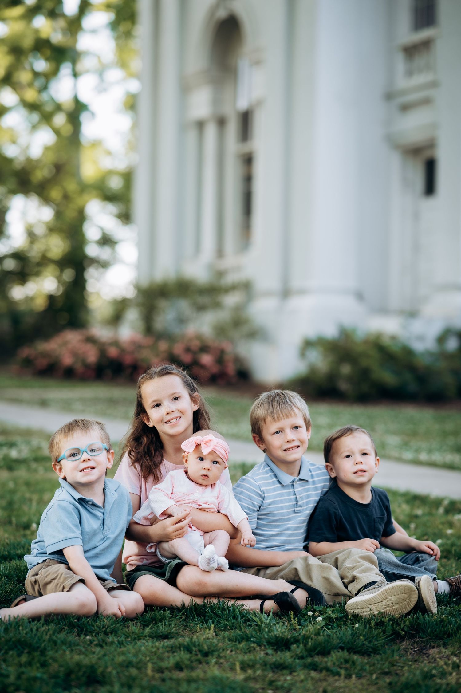 Four young children and a baby sitting on grass in front of a white building for a family portrait.