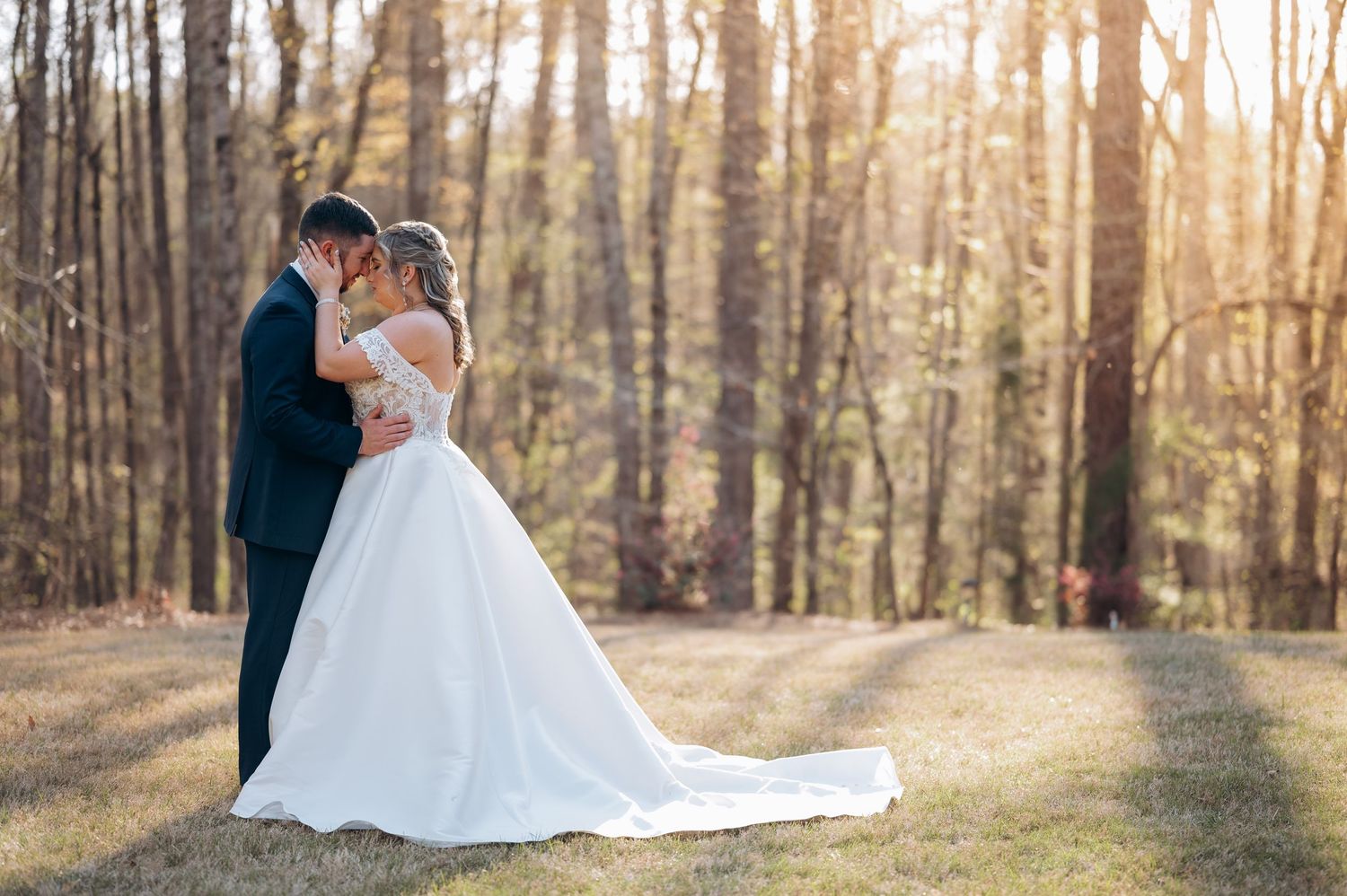 Wedding couple strolls through woods at sunset with dress trailing behind.