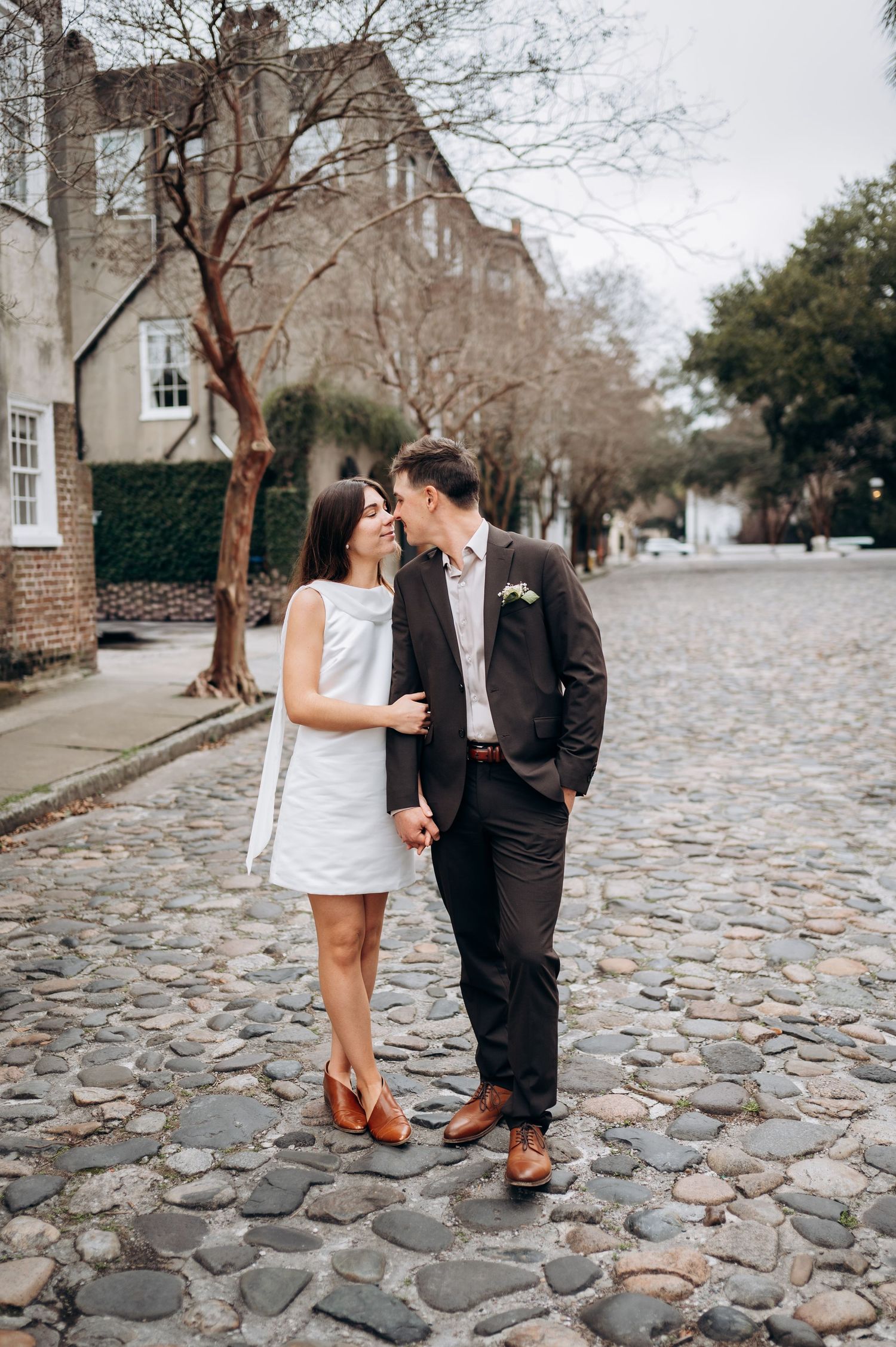 Newlyweds sharing a kiss on historic cobblestone street, bride in white mini dress and groom in dark suit.
