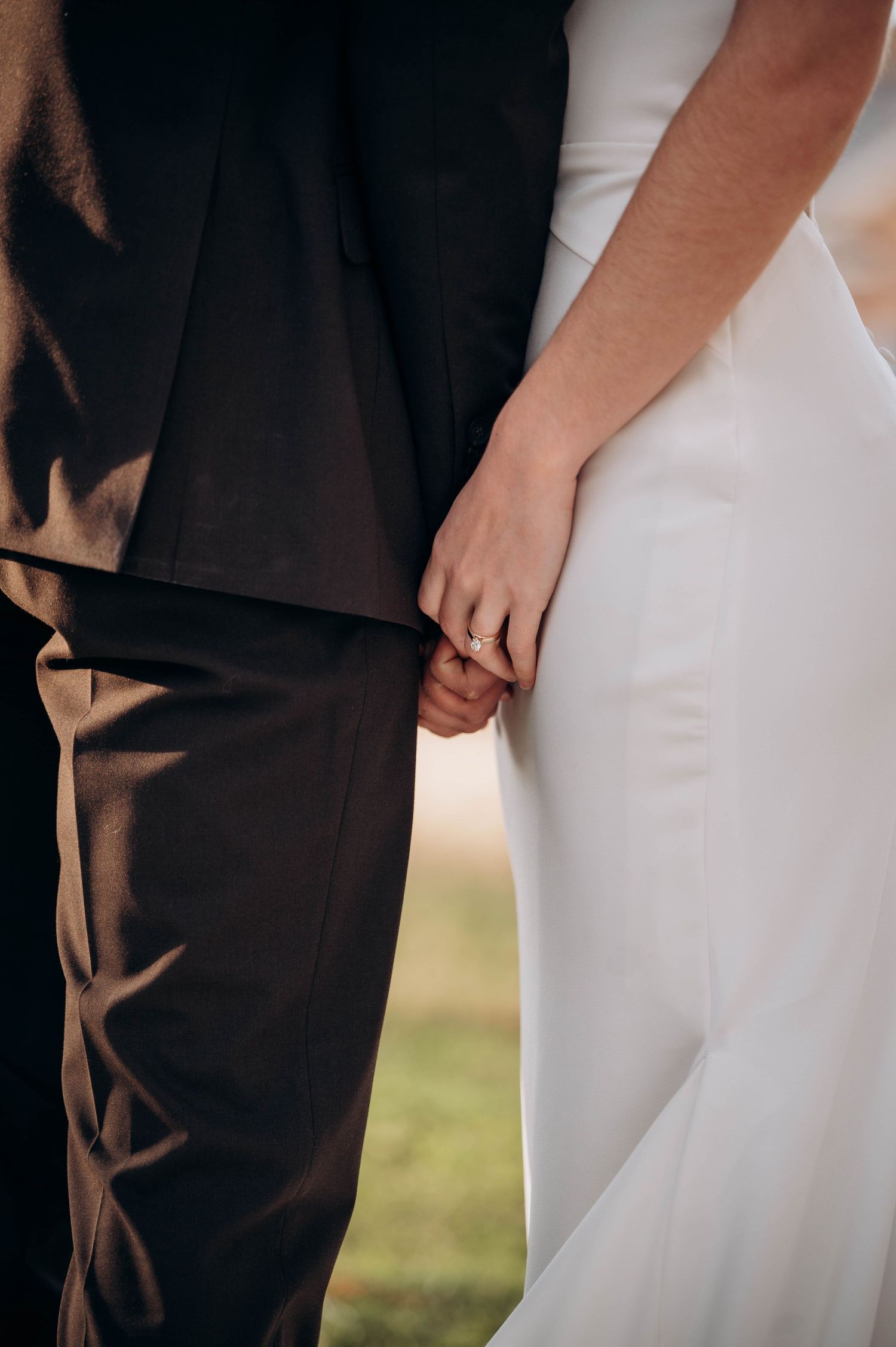 Close-up of bride and groom holding hands, showing wedding ring, bride in white gown and groom in dark suit.