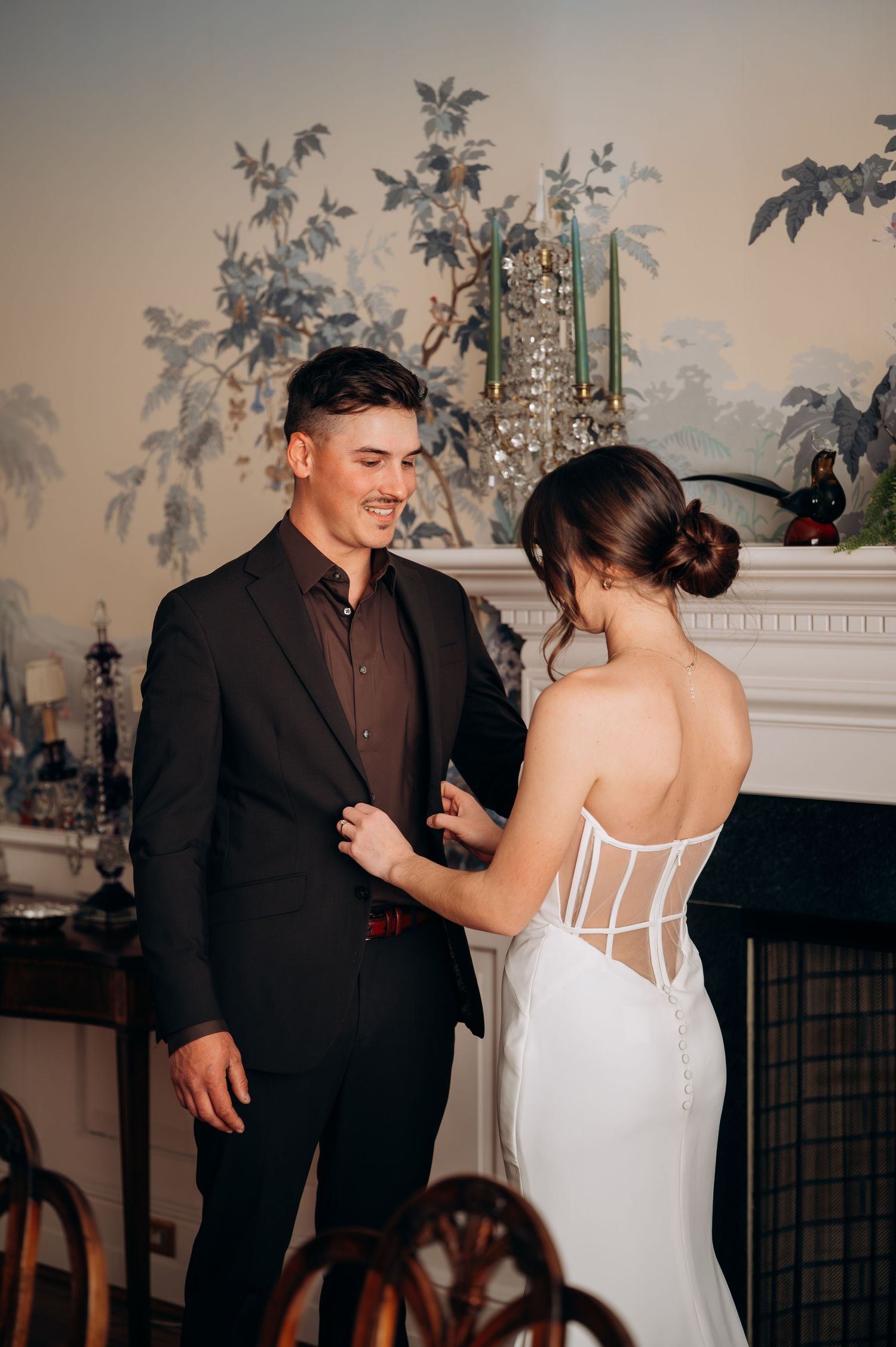 Groom smiling as bride adjusts his dark suit lapel in front of elegant fireplace mantel.