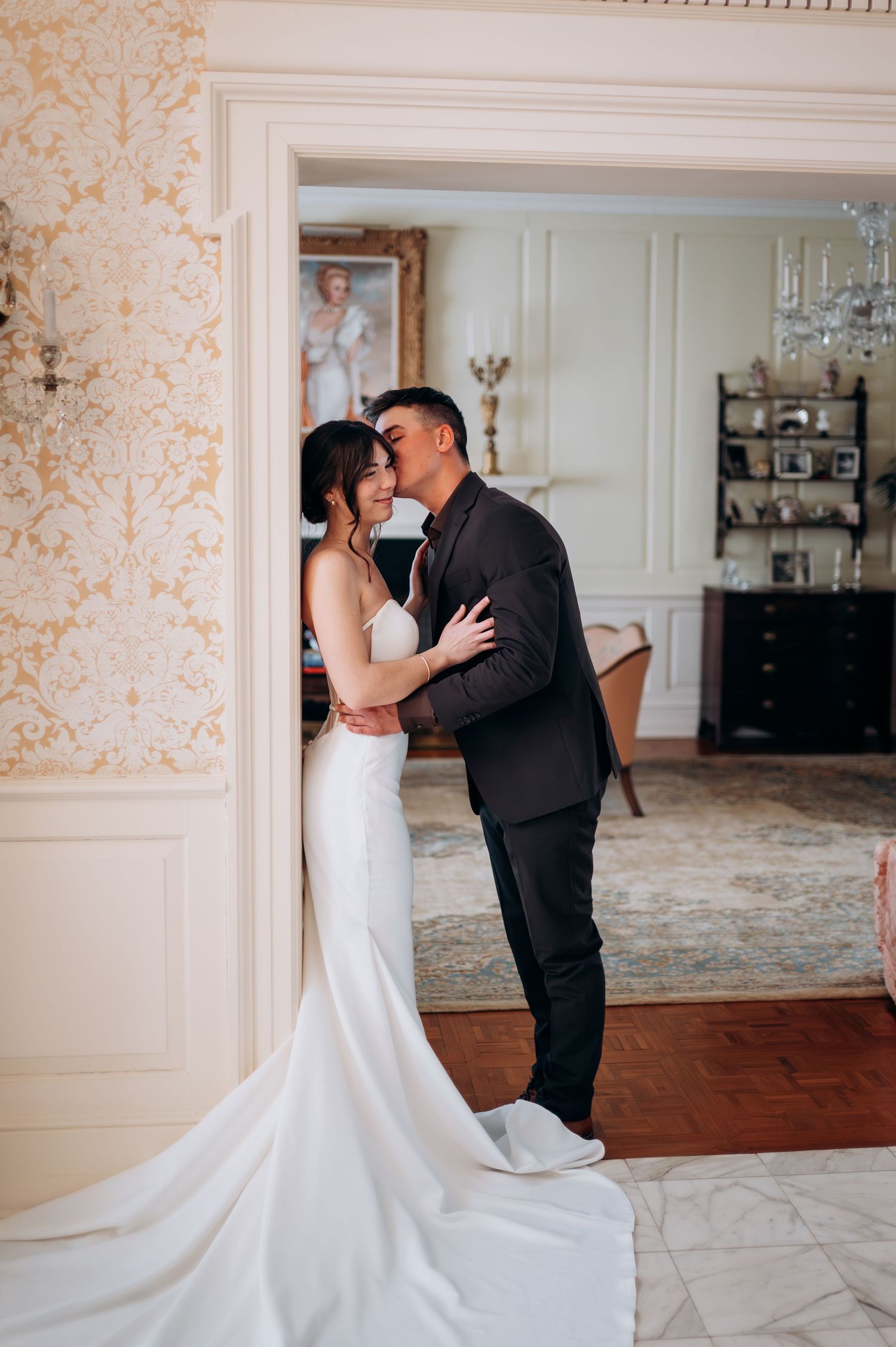 Bride and groom share a romantic embrace in a doorway of an elegant room with gold wallpaper.