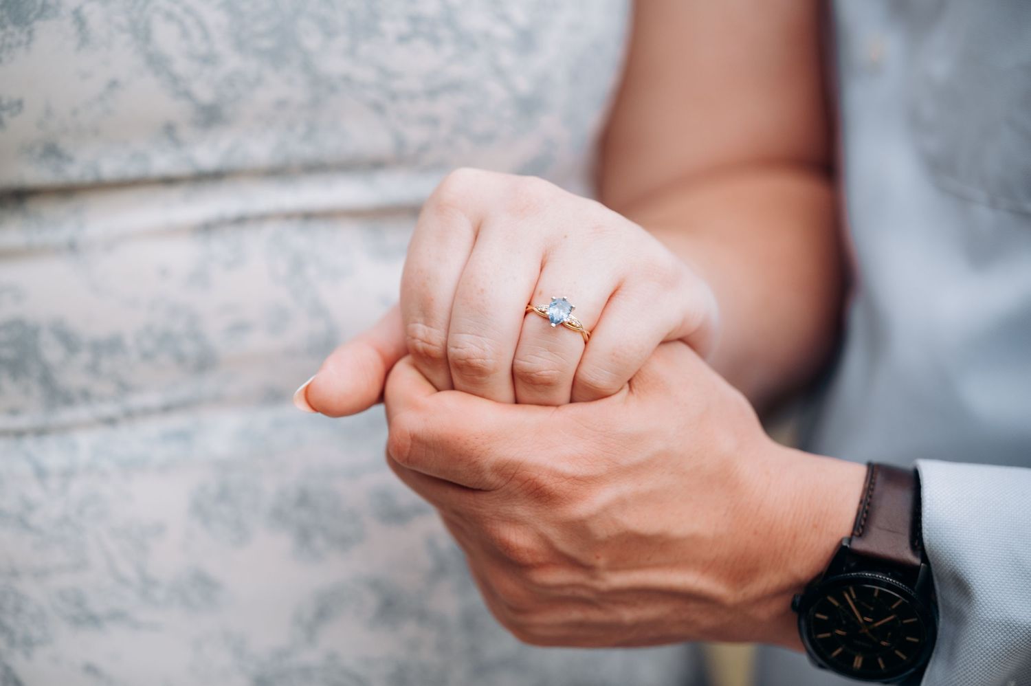 Close-up of womans hand wearing diamond engagement ring held by mans hand with watch.