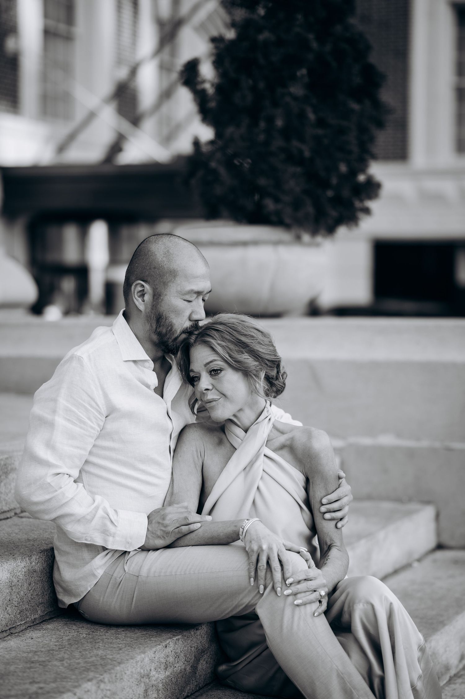 Black and white photo of a romantic couple sitting on steps, embracing tenderly in an urban outdoor setting.
