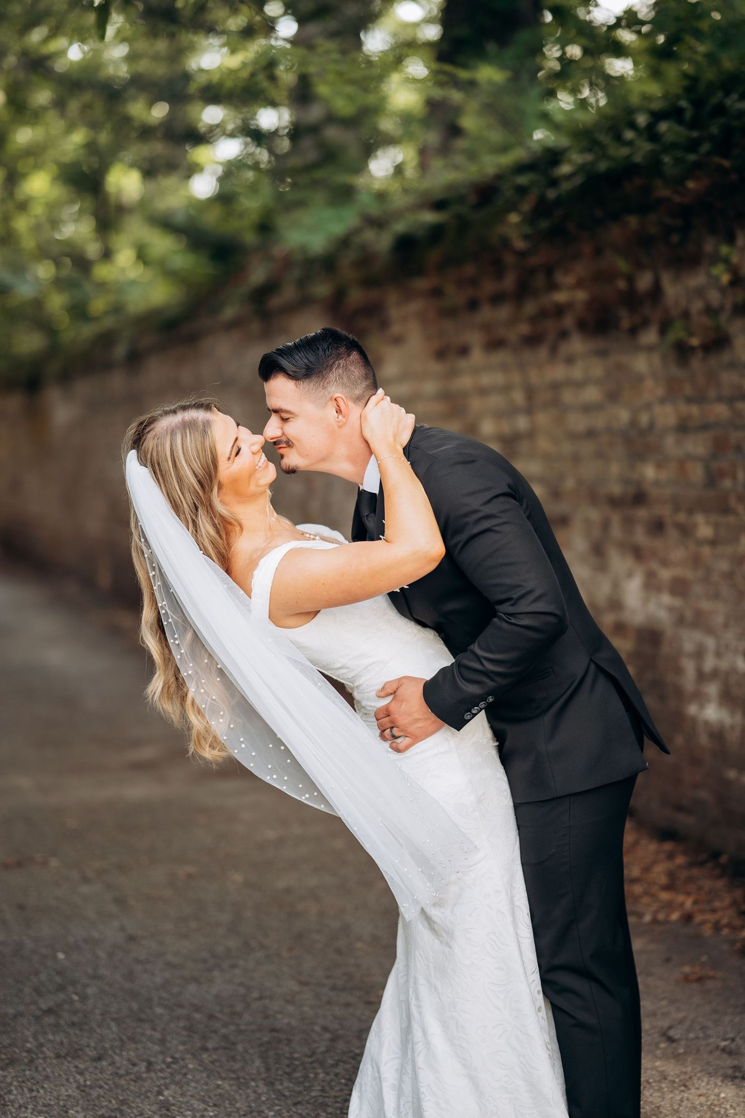Groom dipping bride in white wedding dress near rustic brick wall outdoors.
