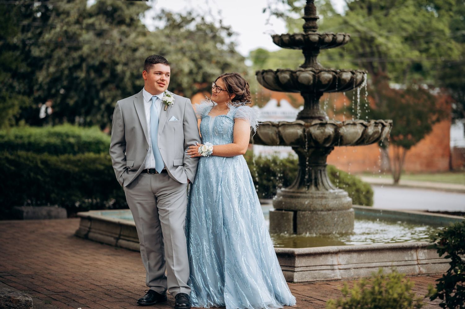 Couple in formal attire posing by a stone fountain, man in gray suit, woman in light blue gown, outdoor garden setting.