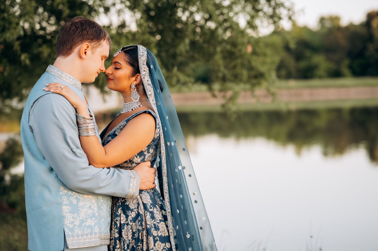 Couple in blue Indian wedding attire sharing romantic moment by serene lake, bride in floral lehenga with dupatta.